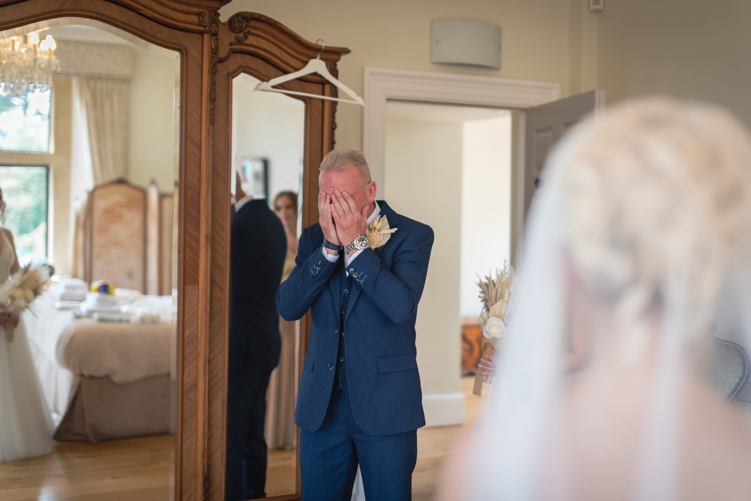 A man in a blue suit with a boutonniere on his lapel is standing in front of a mirror, covering his face with both hands, during a first look with daughter at Old Down Estate in Bristol