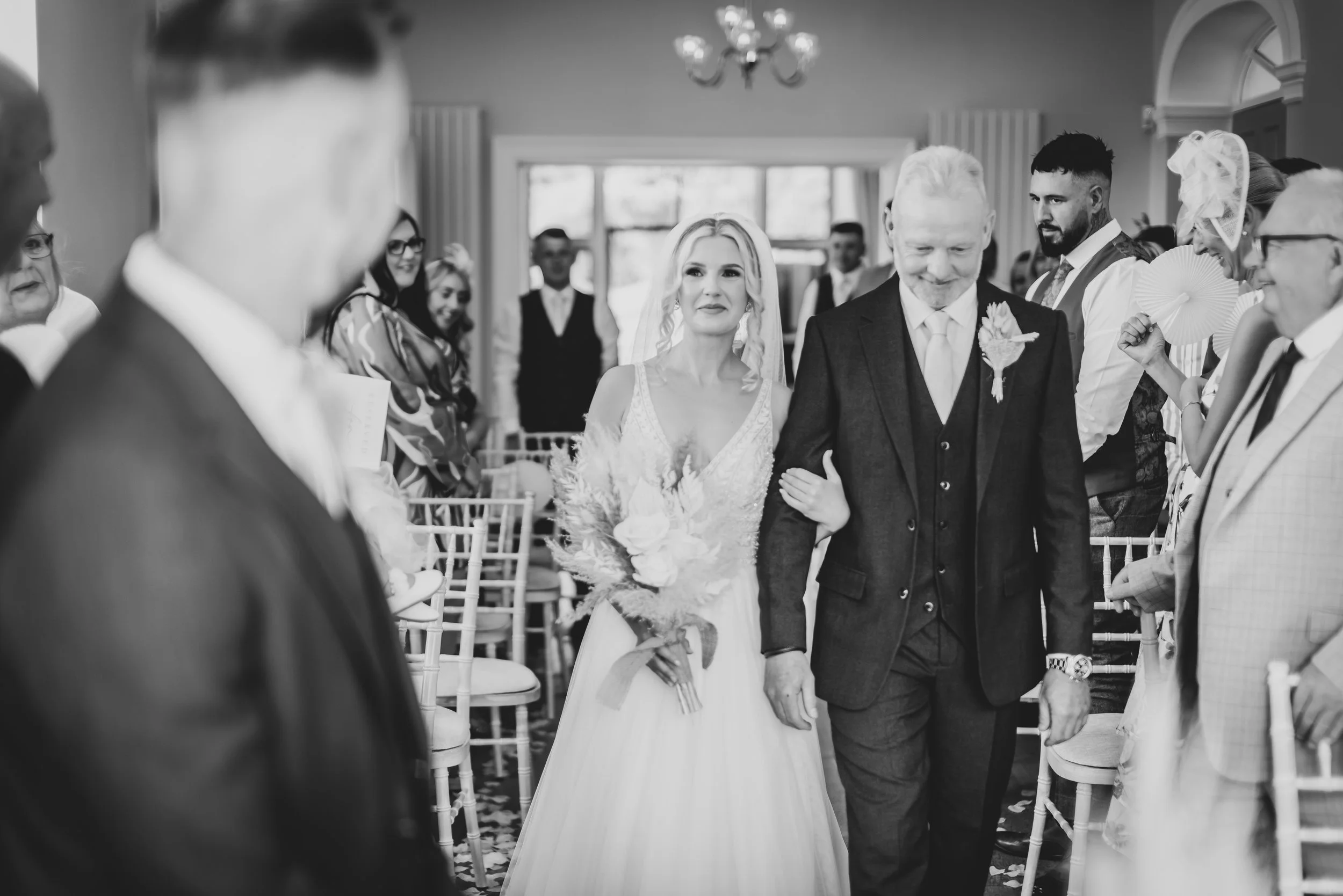 Black and white photo of a wedding ceremony scene, with a bride in a wedding dress being escorted down the aisle by a man in a suit, surrounded by seated and standing guests at Old Down Estate captured by Bristol Wedding Photographer