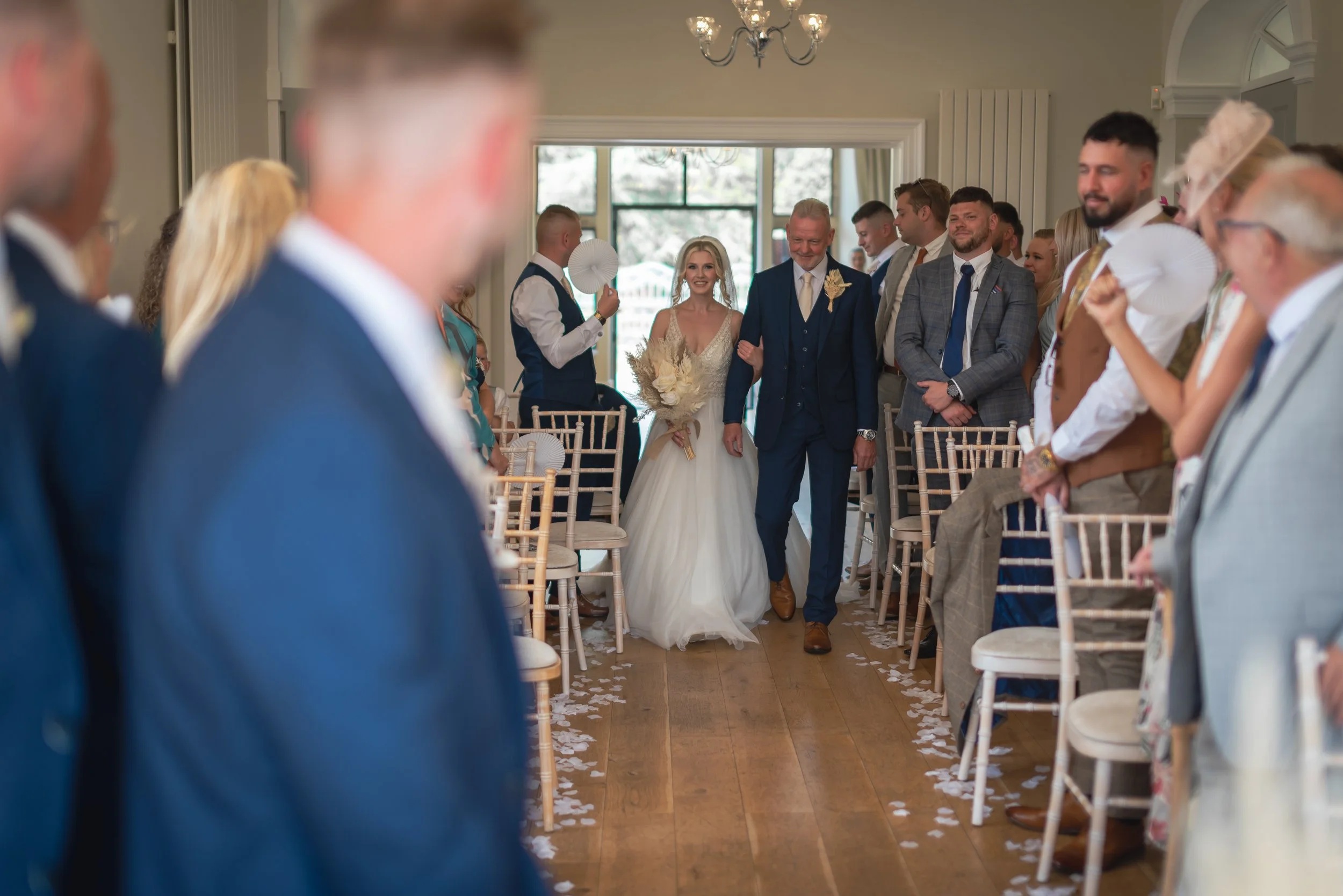 A bride walking down the aisle with her father at a wedding ceremony, surrounded by seated guests in a  decorated Old Down Estate captured by Bristol Wedding Photographer Mike Greenhill
