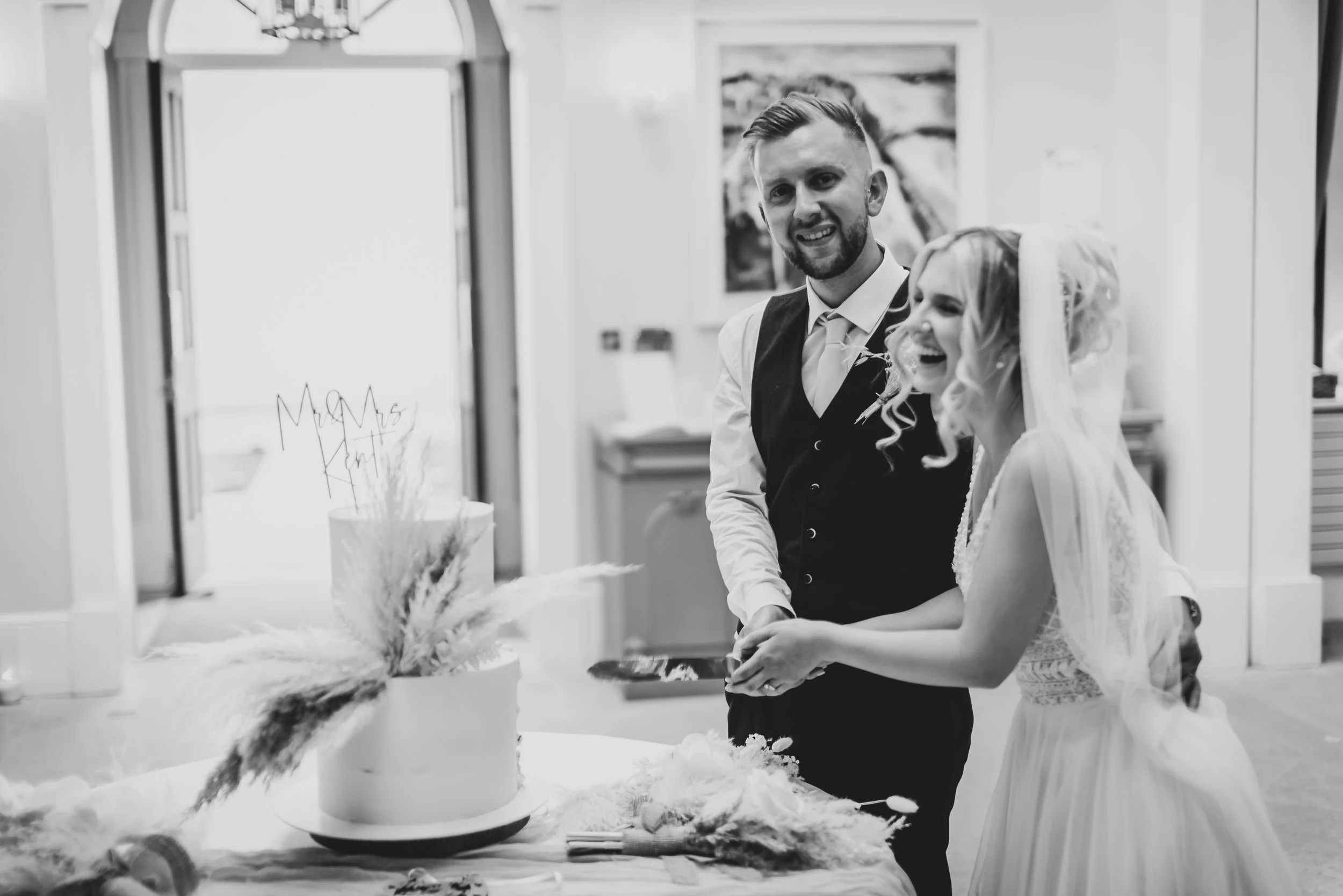 A couple at their wedding, the groom in a vest and tie, and the bride in a wedding dress with veil, both smiling and holding hands as they cut the cake taken by Old Down Estate Wedding Photographer.