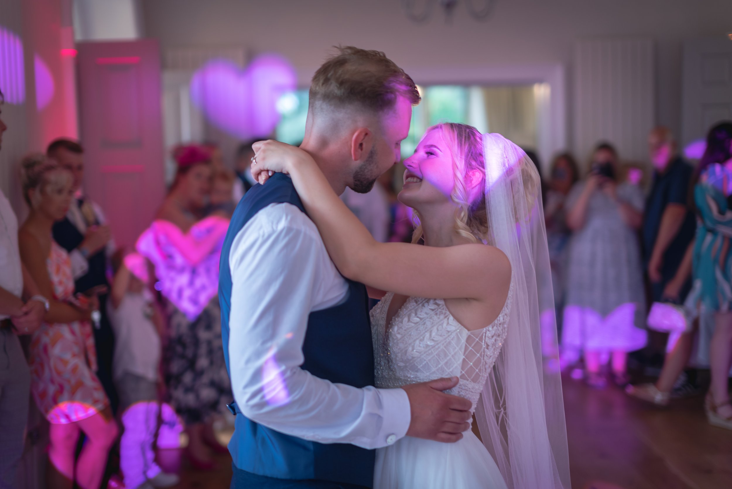 A bride and groom share a dance at their wedding reception, gazing into each other's eyes with smiles, surrounded by wedding guests in a decorated venue with pink lights captured by the Old Down Estate Wedding Photographer.