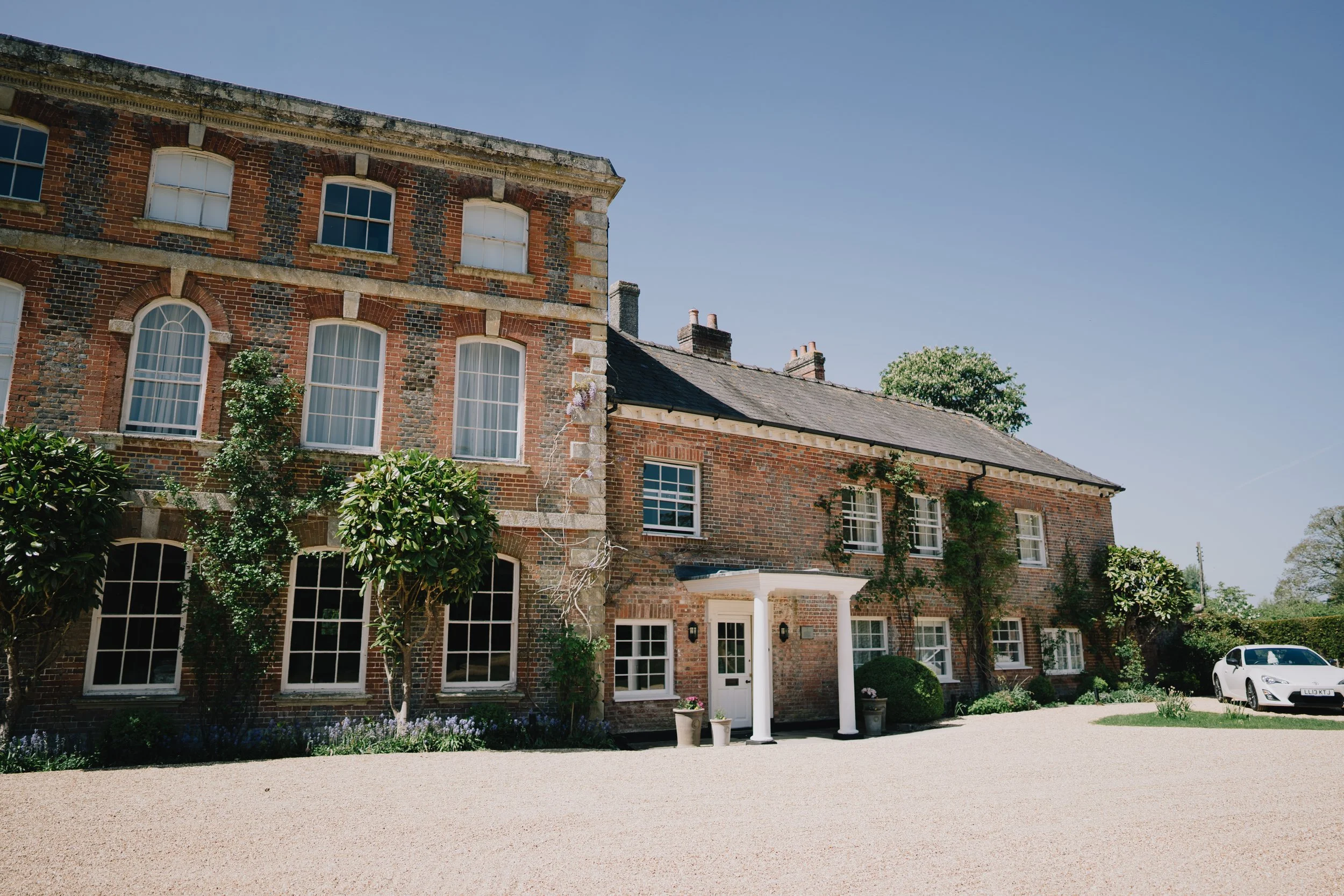Large brick house with white trim, multiple windows, and a driveway with a white car parked on the right. Clear blue sky at Syrencot