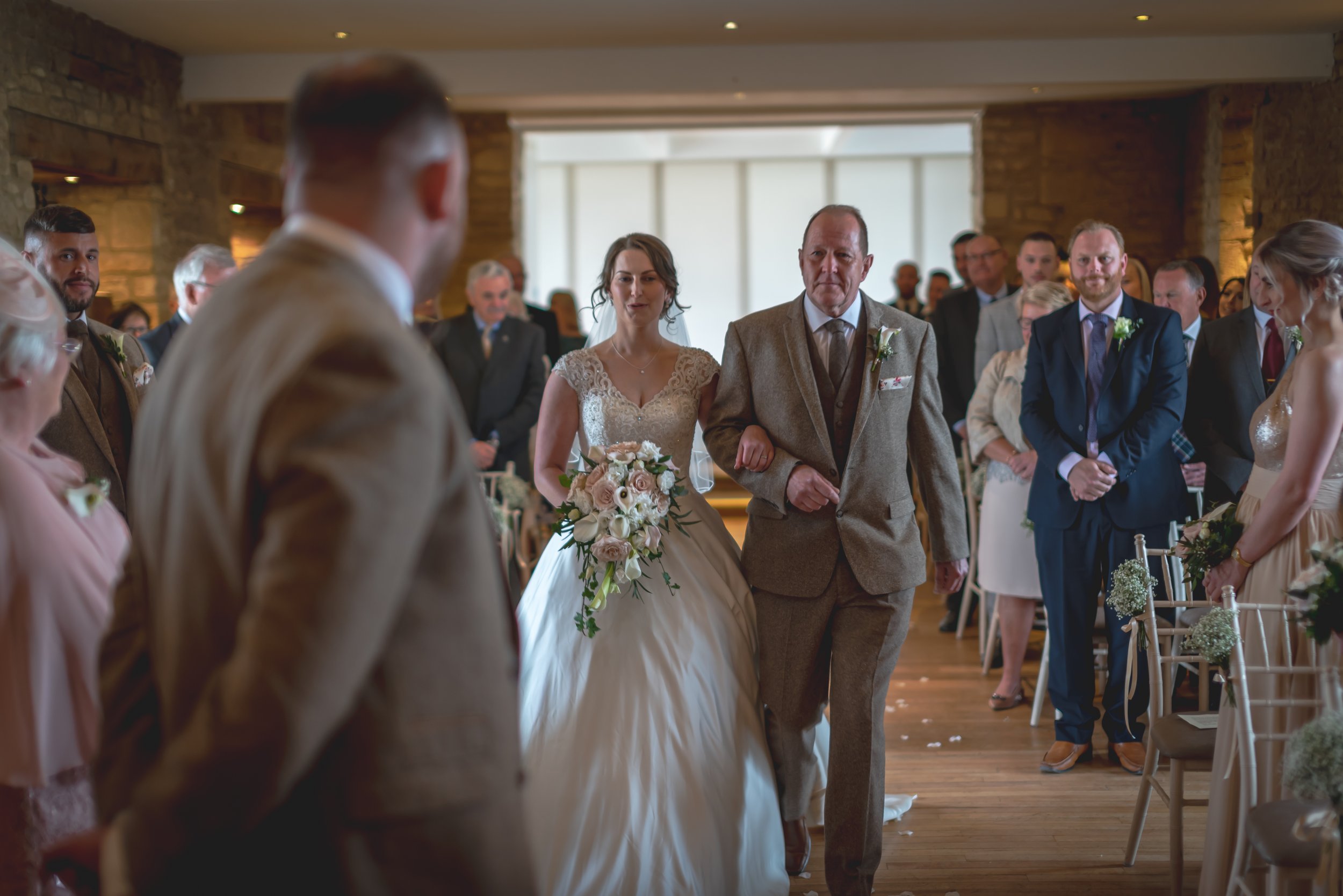 Bridal procession at a wedding ceremony with a bride walking down the aisle escorted by an older man, surrounded by guests in a rustic indoor setting, Greay Tythe Barntaken by Bristol Wedding Photographer