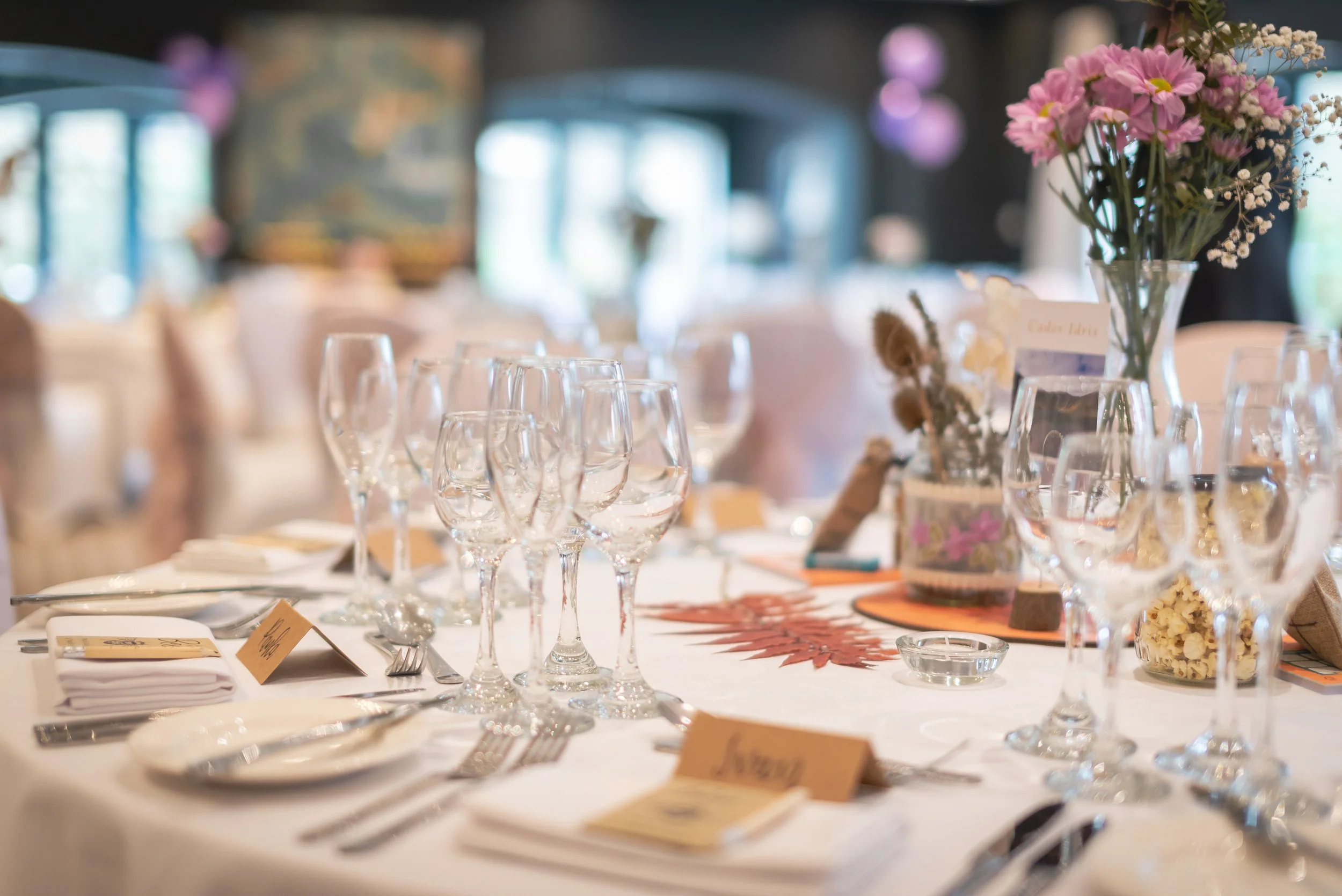 Elegant banquet table set with multiple wine glasses, plates, silverware, and folded napkins, decorated with floral arrangements and place cards, in a well-lit Manor House in Moreton