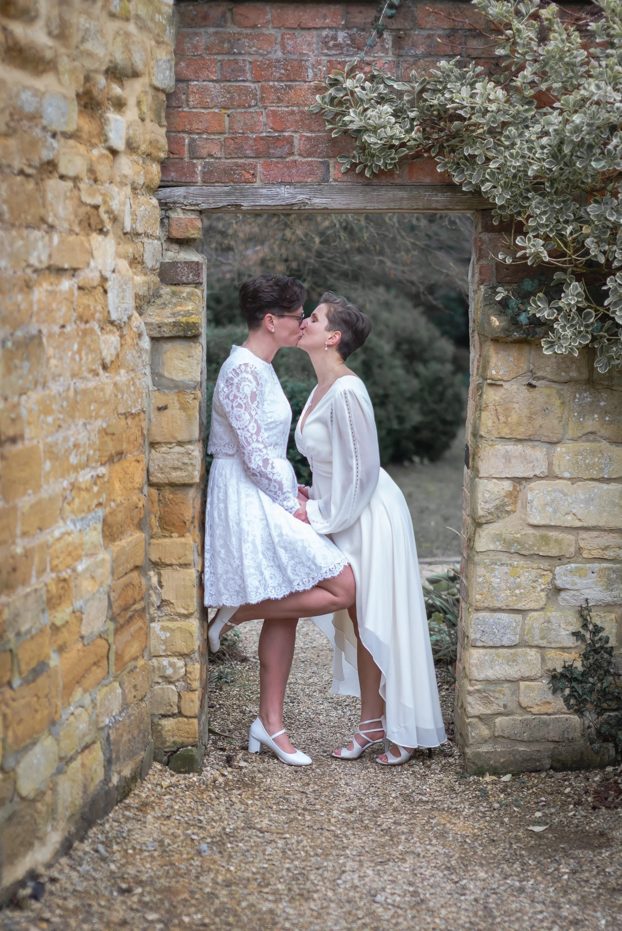 Two women in white dresses kissing in a garden archway with brick and stone walls at Manor House in Moreton