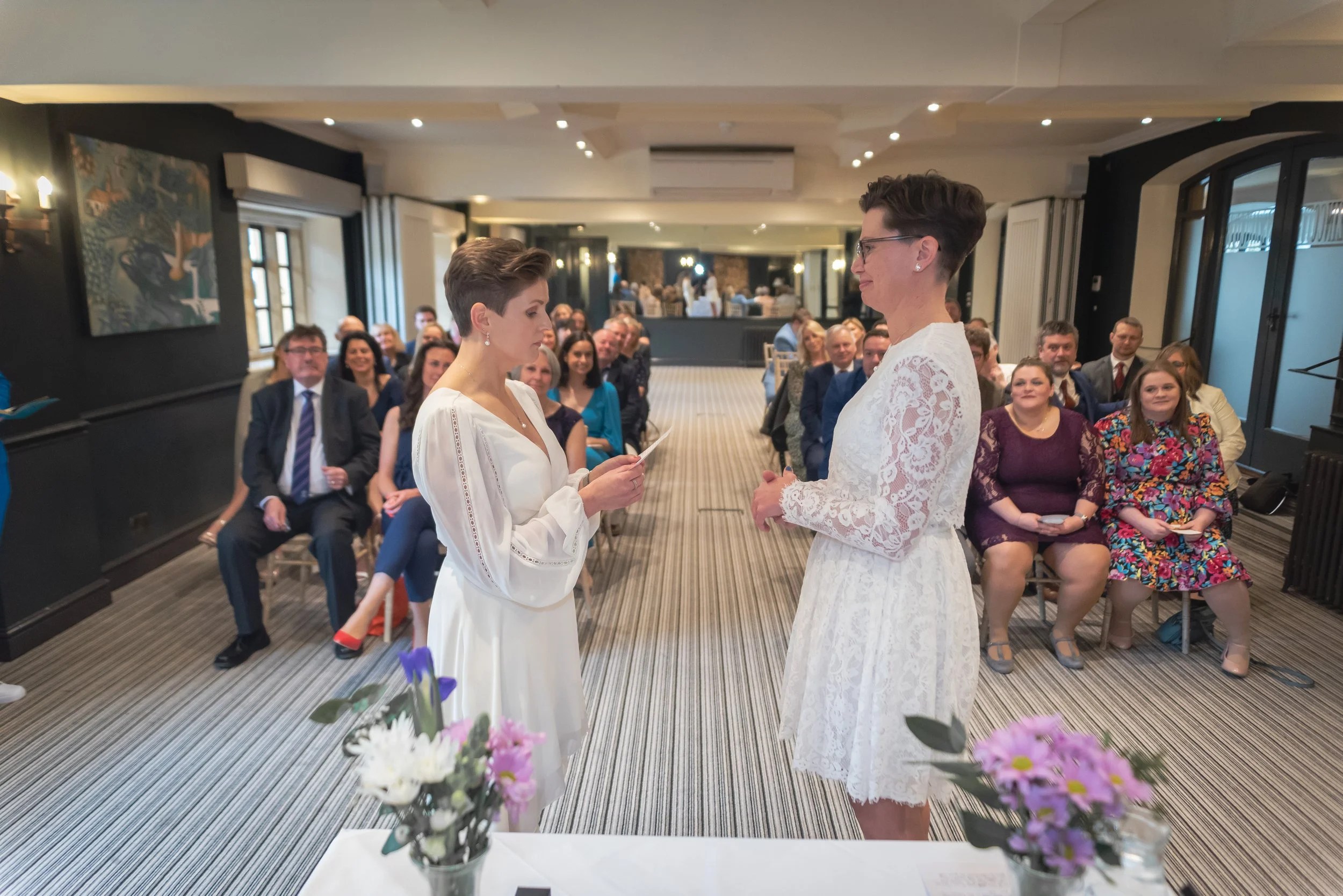 Cotswolds Photographer captures two women in white dresses exchanging vows at a wedding ceremony at Manor House, Moreton in Marsh in a decorated indoor venue with seated guests watching