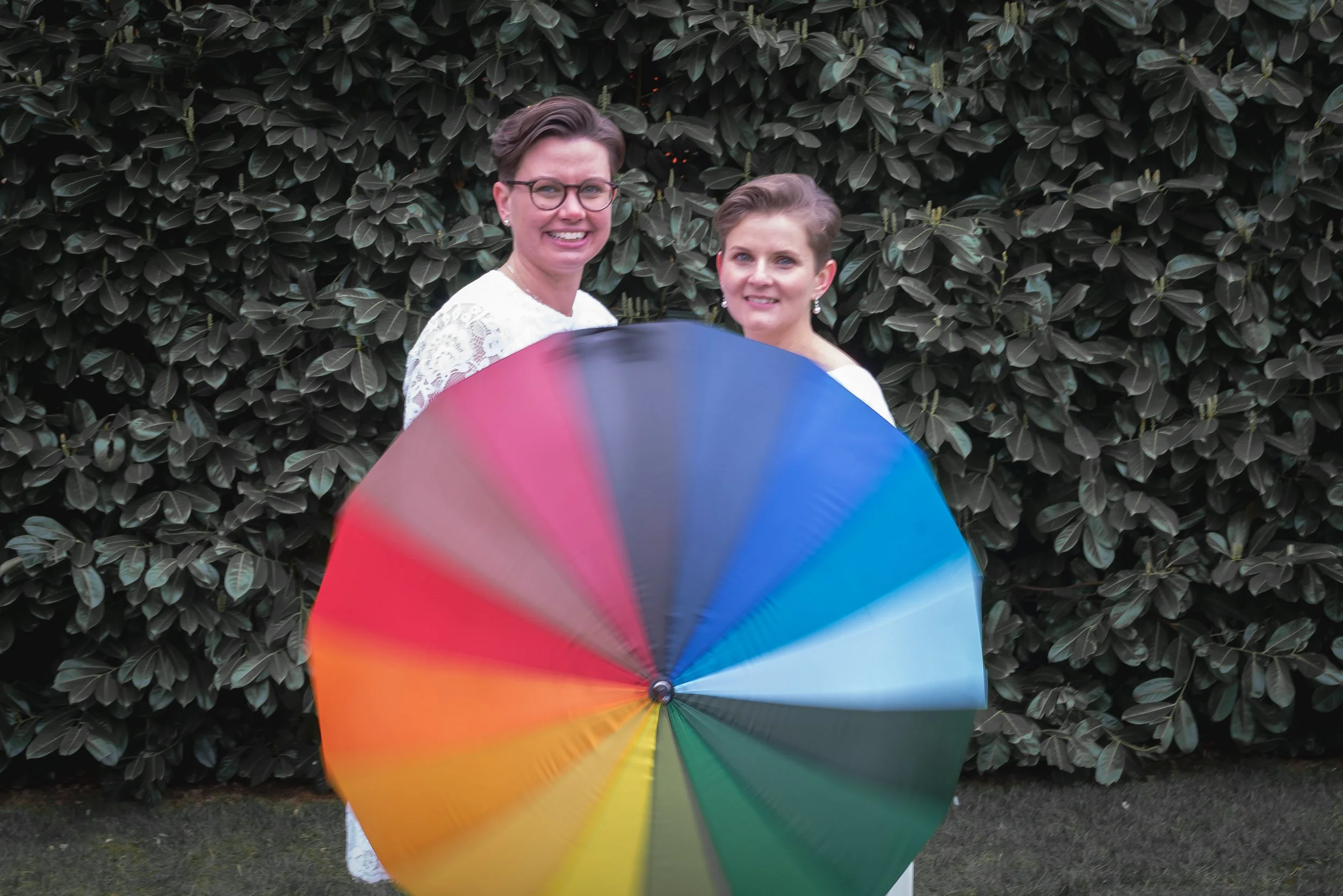 Wedding Photographer captures lgbtqia+ women standing outdoors in front of a dense leafy hedge, holding a large multicolored pride rainbow umbrella, smiling at the camera.