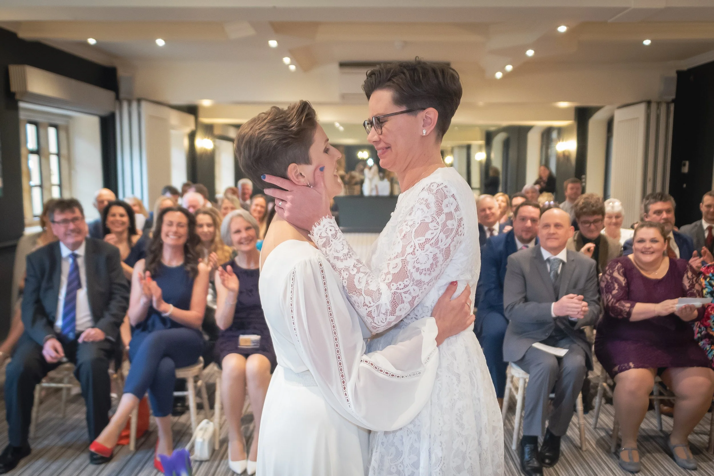 Two women embracing and smiling at each other during a wedding ceremony. They are surrounded by an applauding seated audience in a decorated indoor venue at Manor House in Moreton.