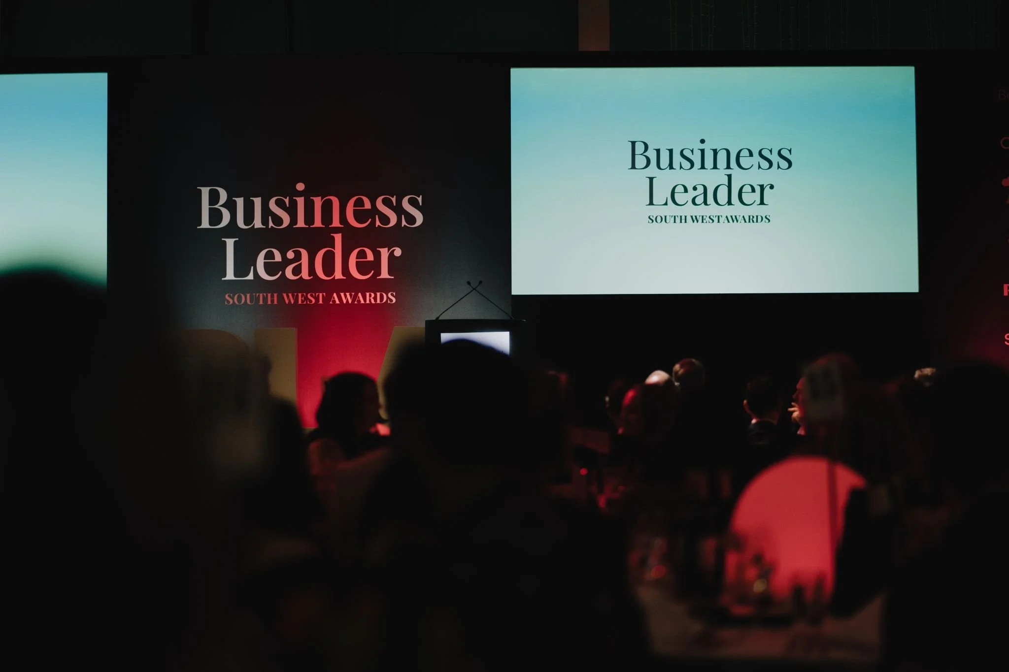 Audience seated at tables during an awards event with two large screens displaying 'Business Leader South West Awards' and similar text.