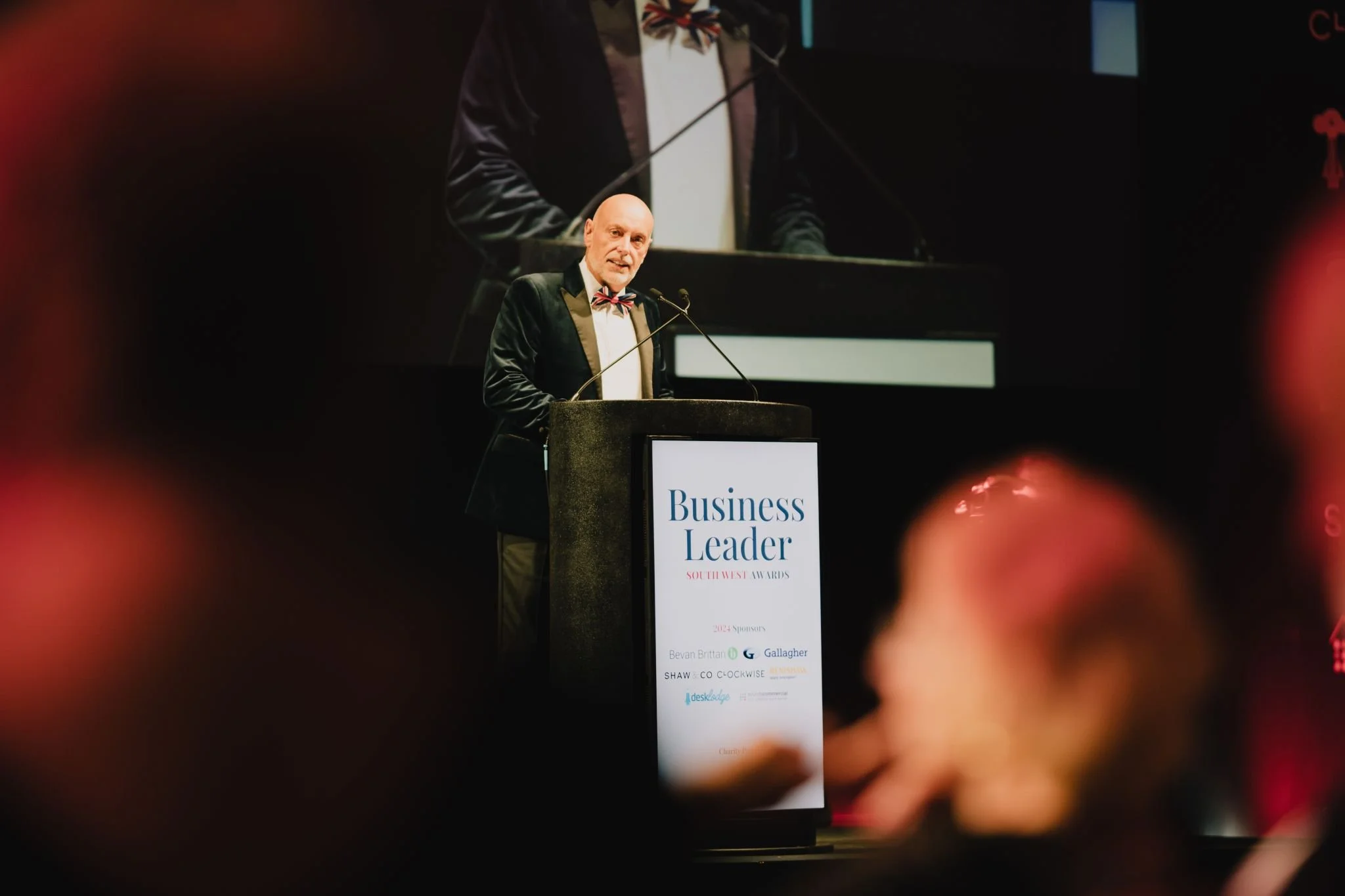 A man in formal attire with a bow tie speaking at a podium during a business awards event, with a large screen behind him displaying his image.
