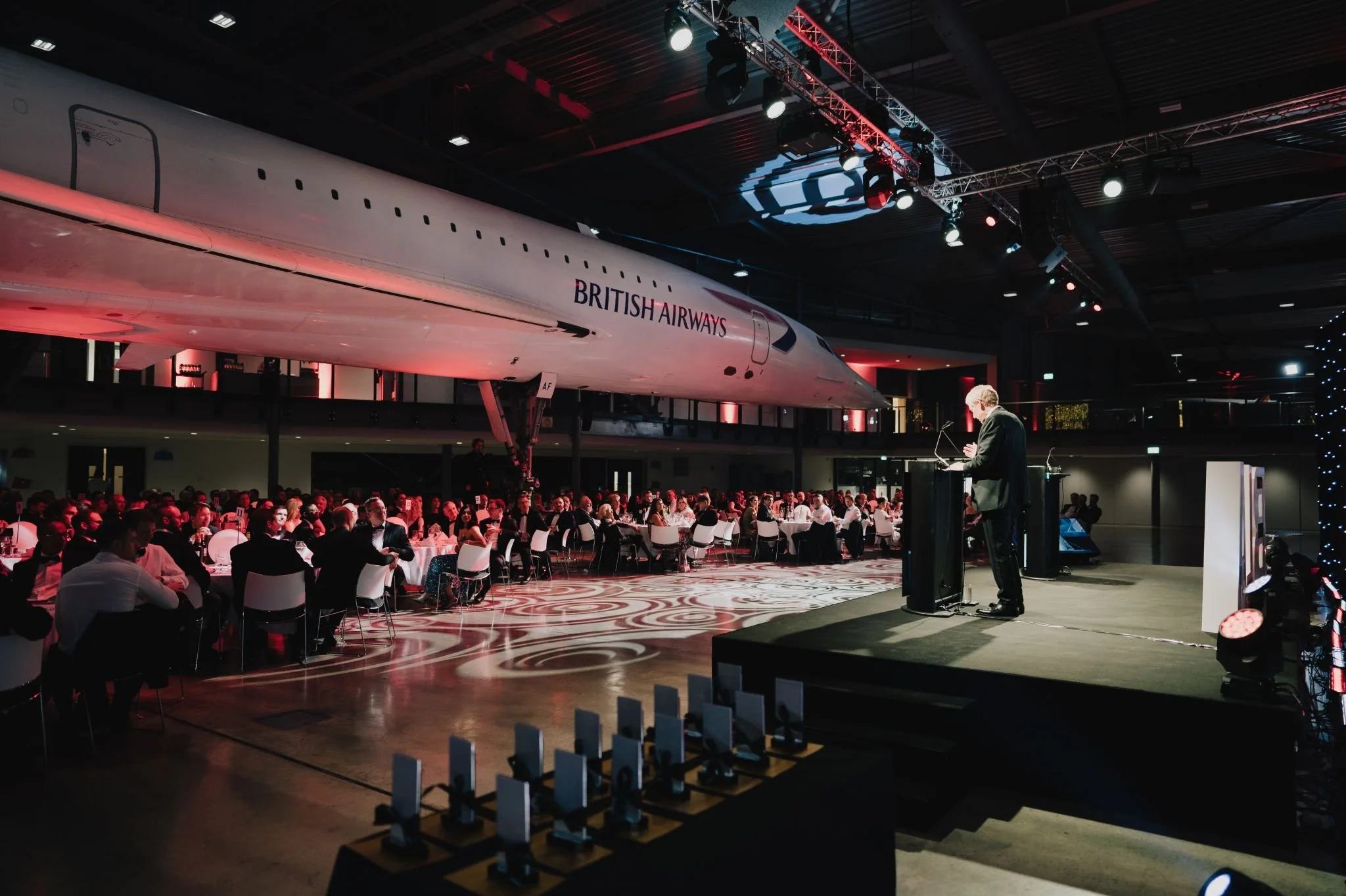 A formal event or dinner with many attendees seated at round tables inside a large hall. An airplane fuselage with 'British Airways' branding is displayed overhead as part of the event's decor. A speaker is standing at a podium on stage, addressing the audience, with stage lighting illuminating the scene.