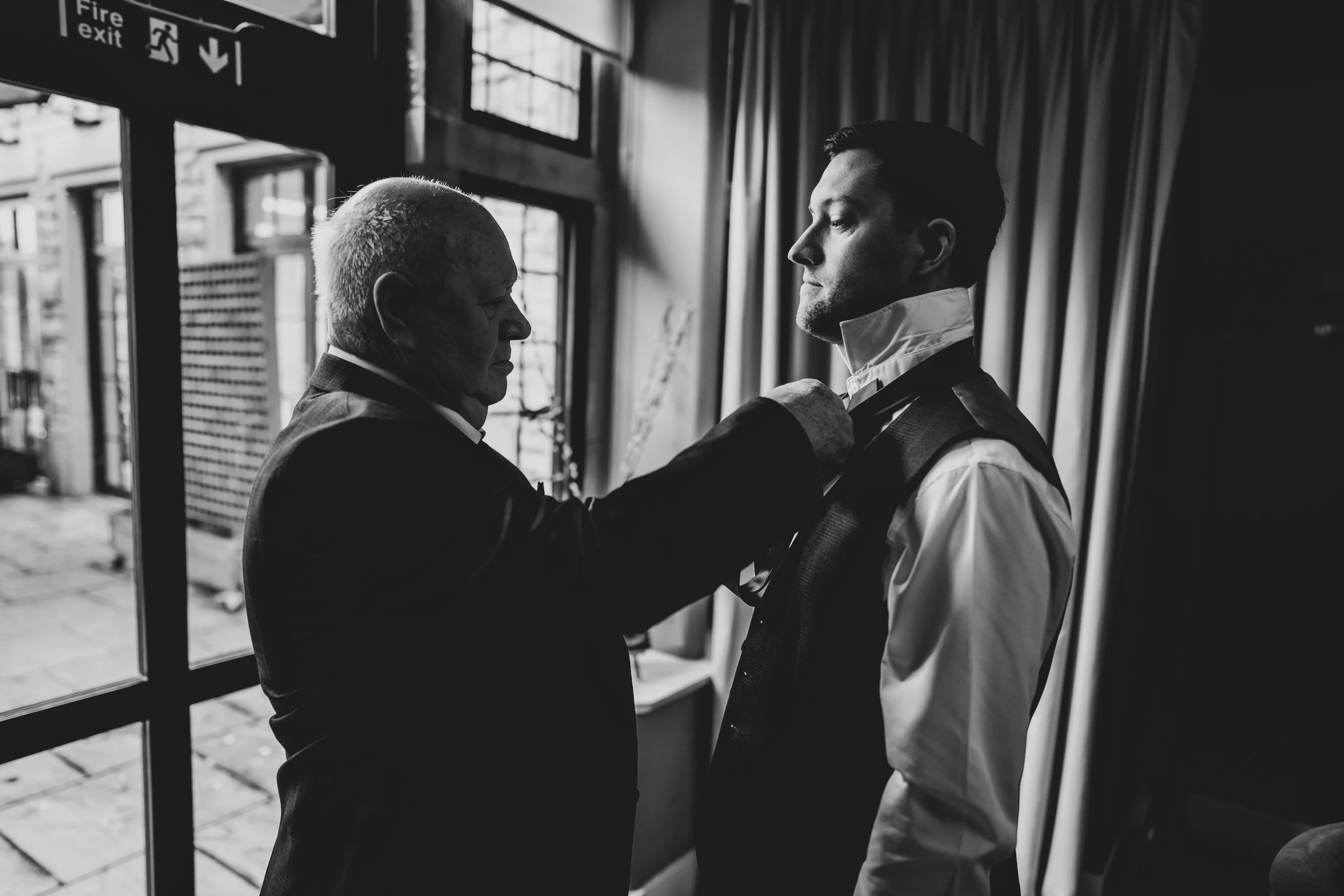 Father adjusting a Grooms bowtie indoors with window in background taken by Wiltshere Wedding Photographer