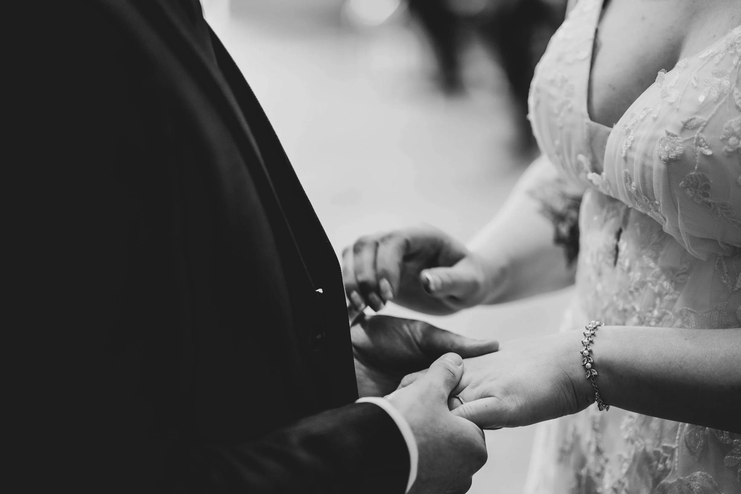A close-up black and white photo of a couple holding hands during their wedding ceremony, with the bride wearing a lace wedding dress and a bracelet, and the groom in a dark suit.