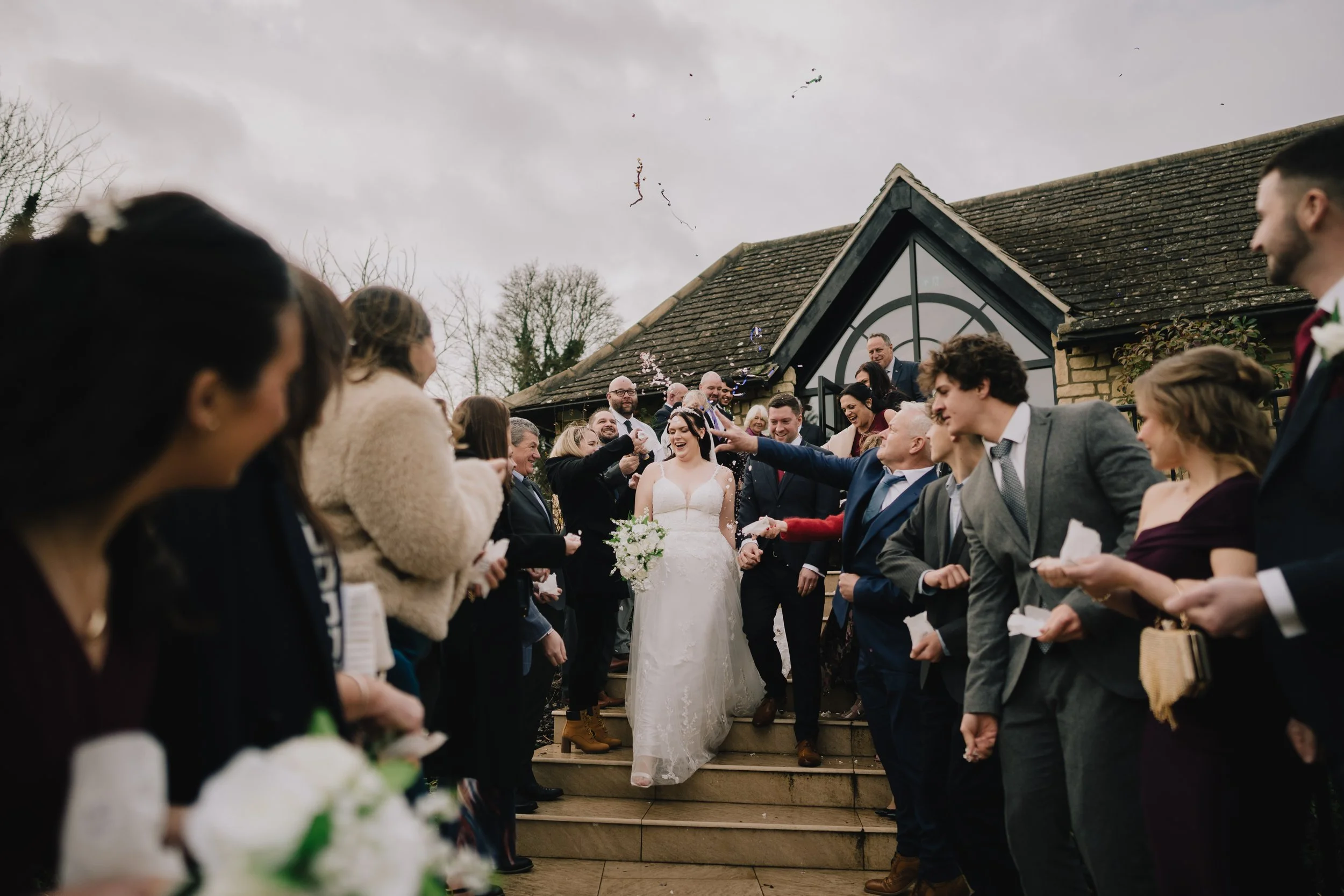 Wedding celebration outside with guests throwing confetti as bride and groom hold hands and smile after their ceremon at Pear Tree taken by Wiltshere Wedding Photographer
