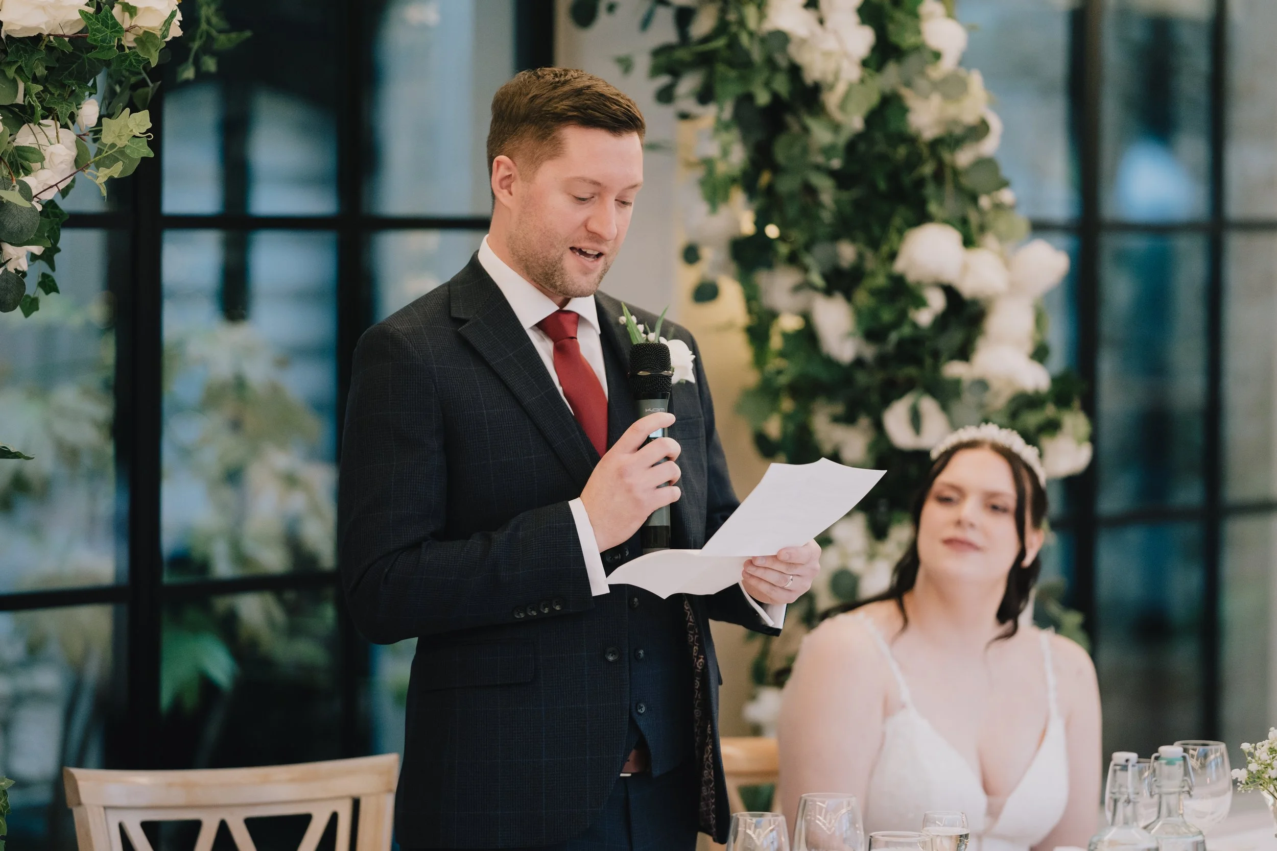 The groom in a dark suit giving a speech at a wedding reception, standing next to the bride in a white dress seated at a decorated table at the Pear Tree during Christmas wedding