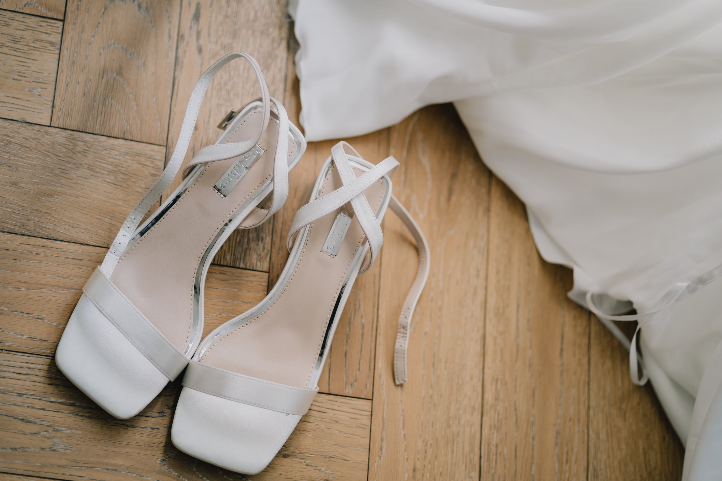 White high-heeled sandals with ankle straps on a wooden floor next to white wedding dress during bridal prep