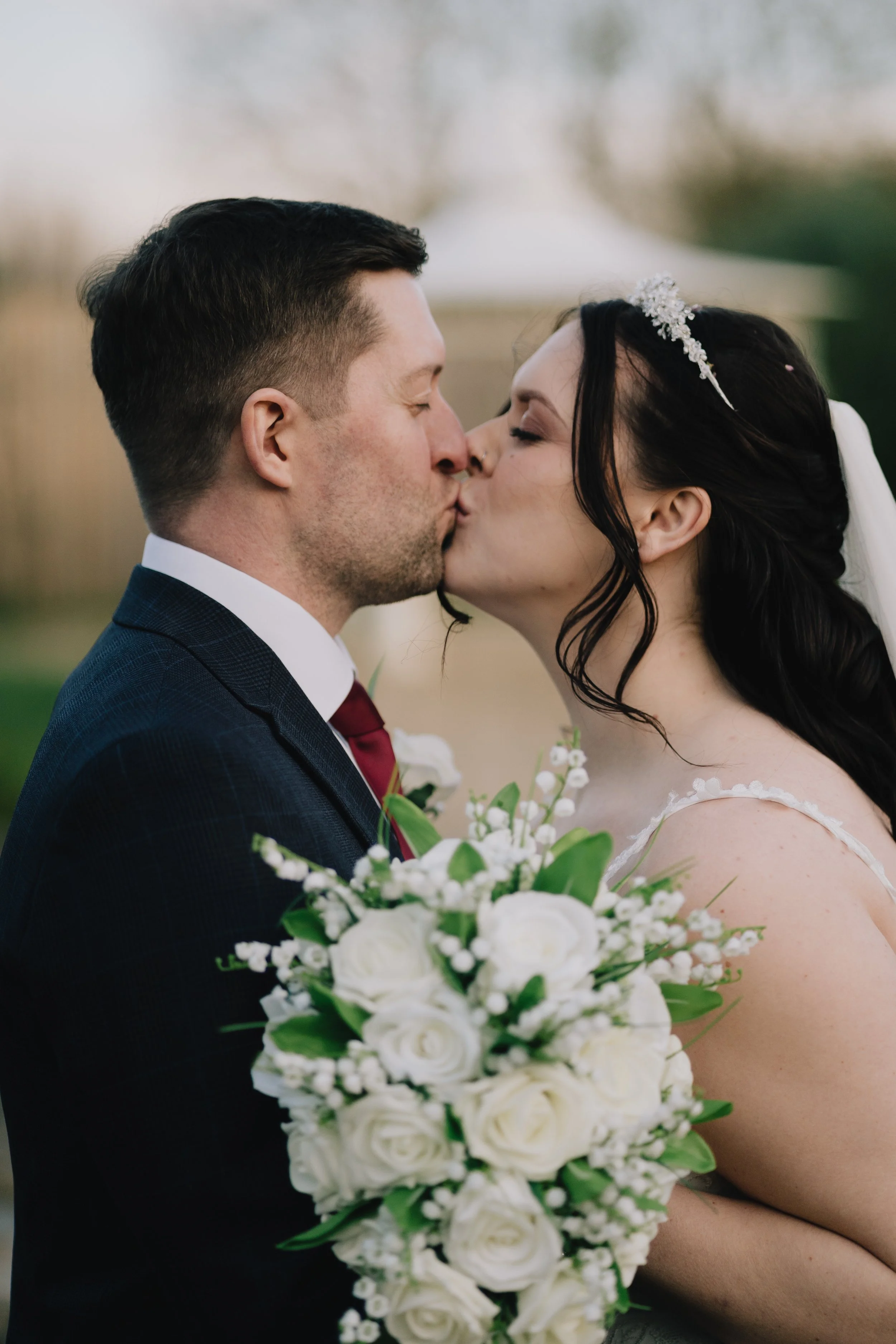 A bride and groom sharing a kiss on their wedding day, with the bride holding a bouquet of white roses and dressed in a wedding gown with a veil and tiara, and the groom in a dark suit with a white shirt and red tie.