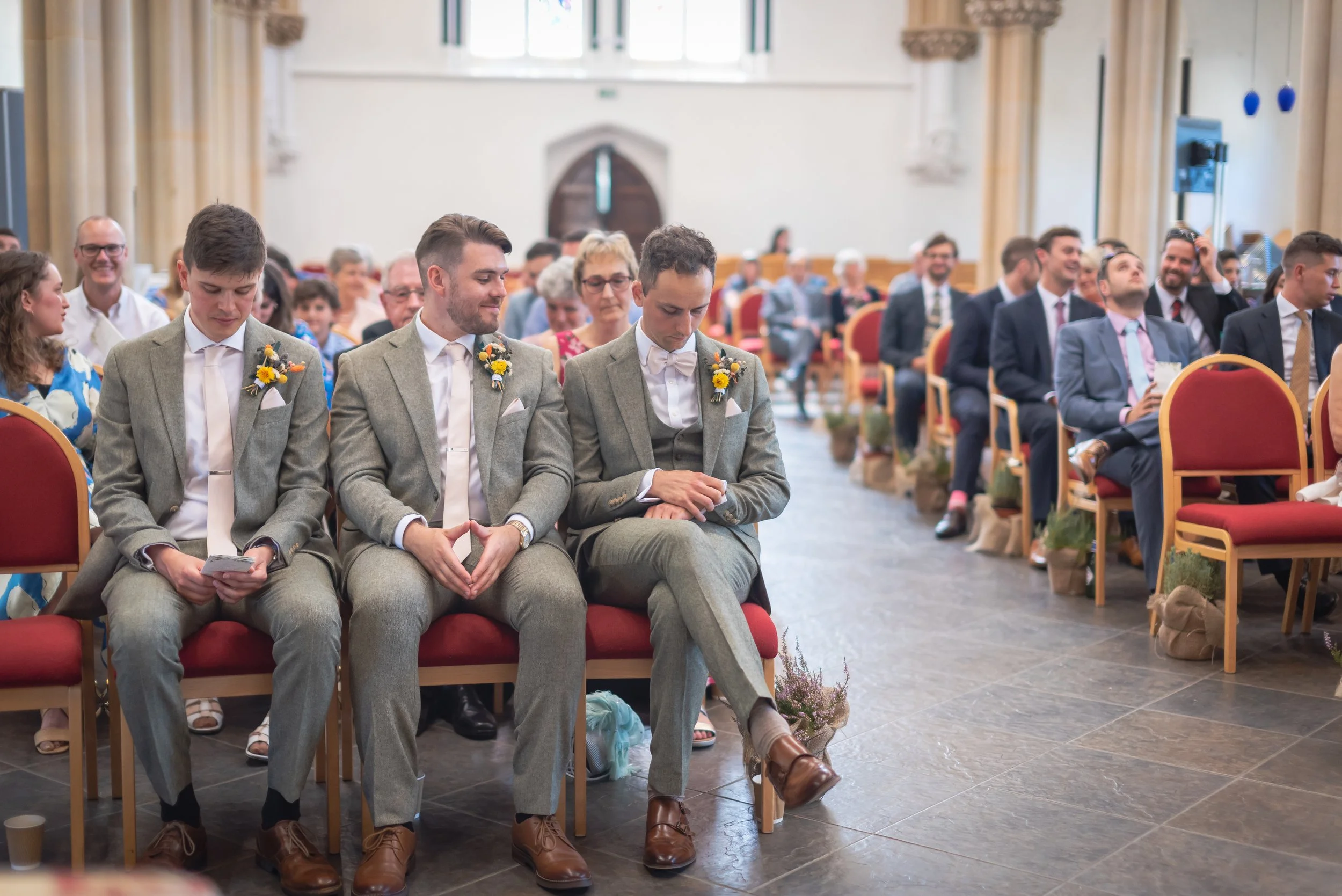 Three young men in gray suits with yellow flower boutonnieres sitting in a church during a wedding ceremony, with guests seated in the background waiting for the bride to arrive