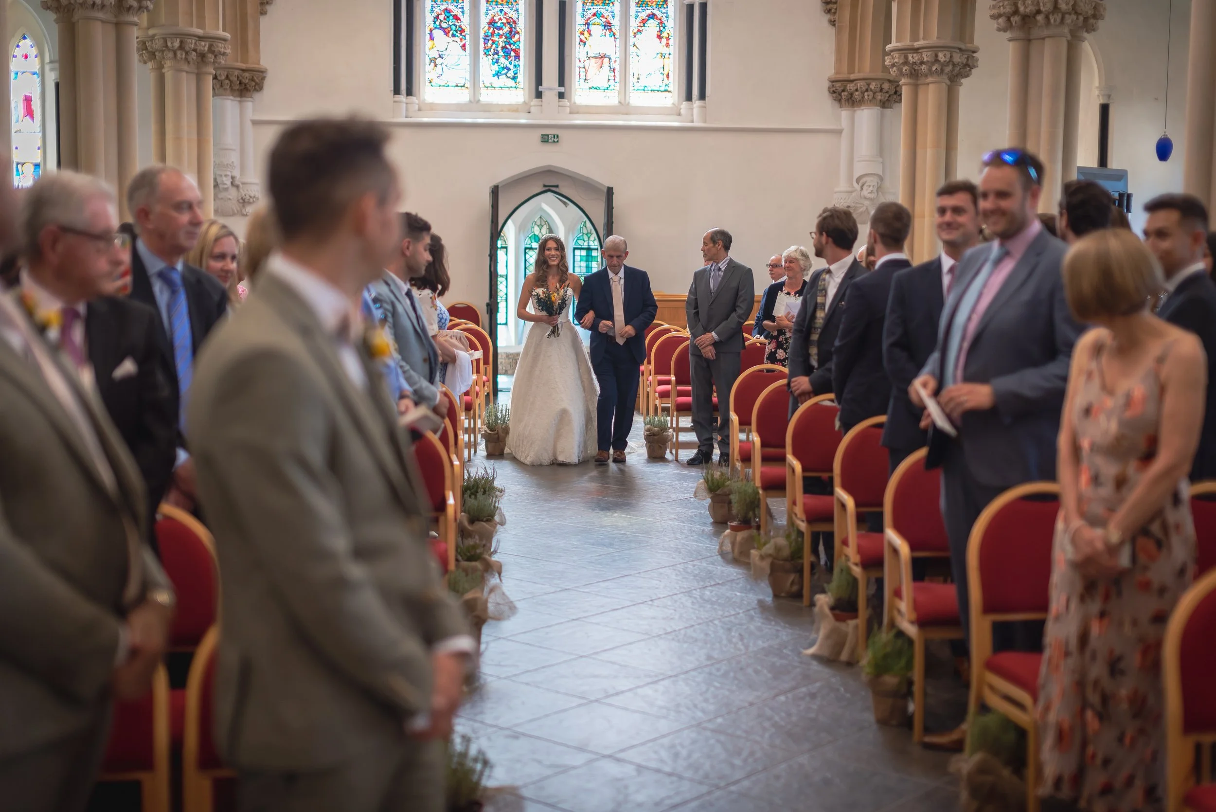 A wedding ceremony inside a church with a bride walking down the aisle escorted by a man, possibly her father, with guests standing on either side of the aisle watching and smiling. The church has stained glass windows and ornate architectural details.