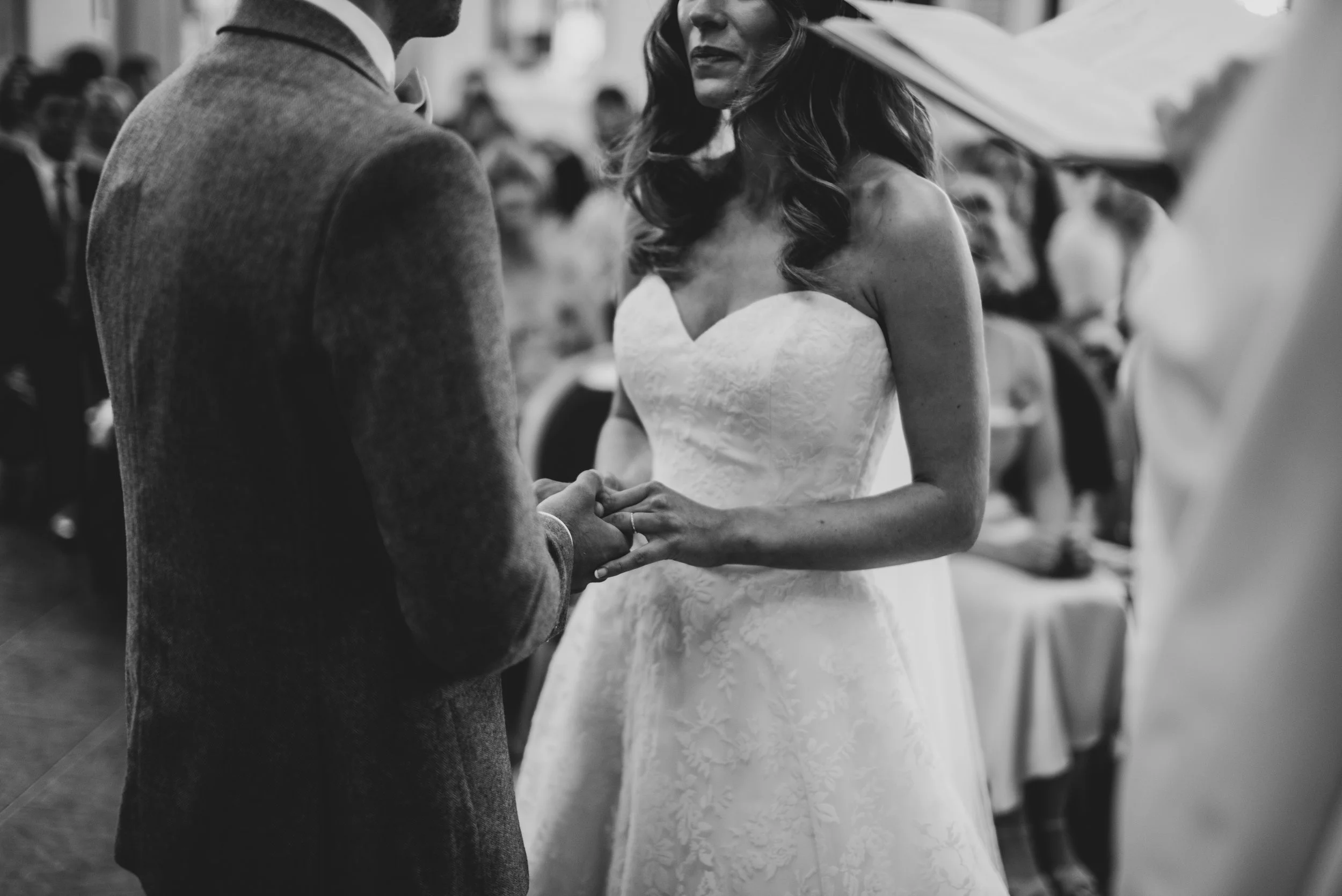 Black and white photograph of a bride and groom exchanging vows, holding hands during a wedding ceremony at an outdoor setting with guests seated in the background.