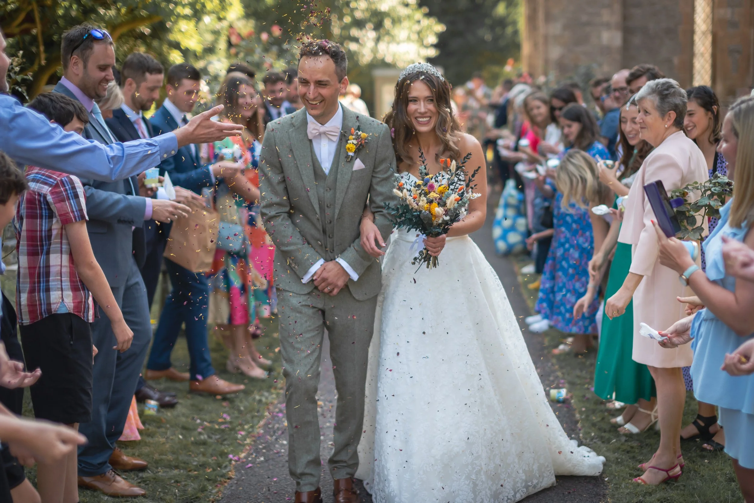 A newlywed couple walking arm-in-arm through a crowd of friends and family during an outdoor wedding celebration, with confetti falling around them coordinated by Bristol wedding photographer.