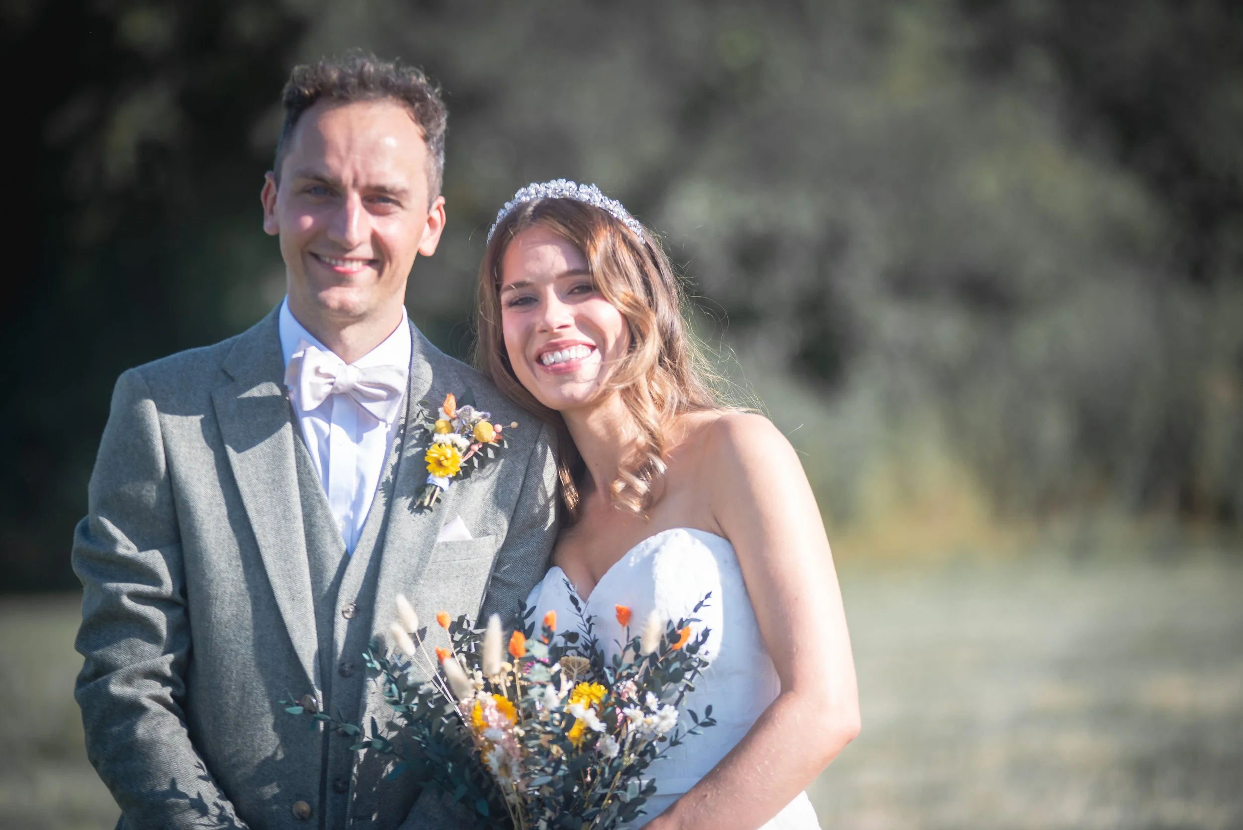 Happy couple on their wedding day outdoors, the groom in a gray suit with a white bow tie and boutonnière, the bride in a strapless white dress with a floral bouquet, both smiling at the camera.