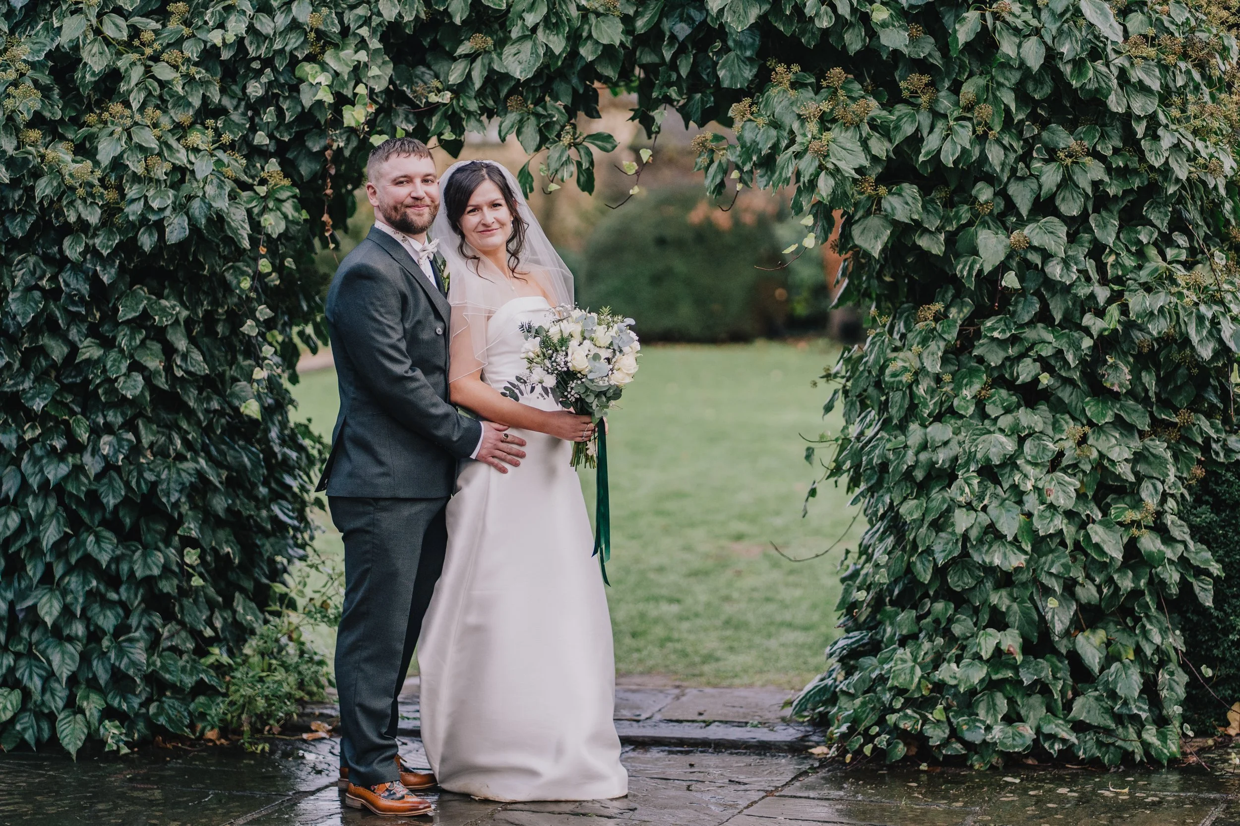 A bride and groom standing together outdoors under an archway of green leafy vines, smiling, with the bride holding a bouquet of white and purple flowers at Manor House Hotel.