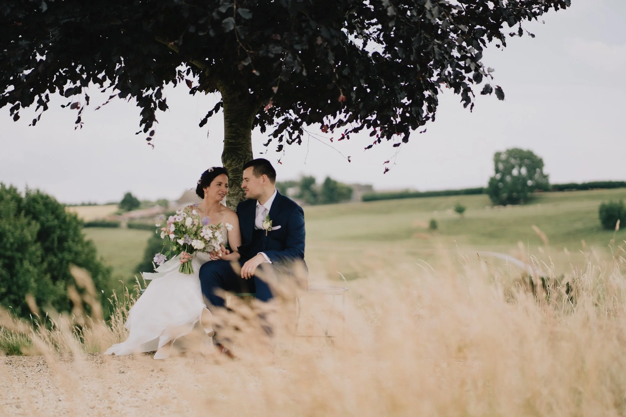 Bride and groom sitting under a tree on a hill at Kingscote Barn, embracing and enjoying a quiet moment on their wedding day