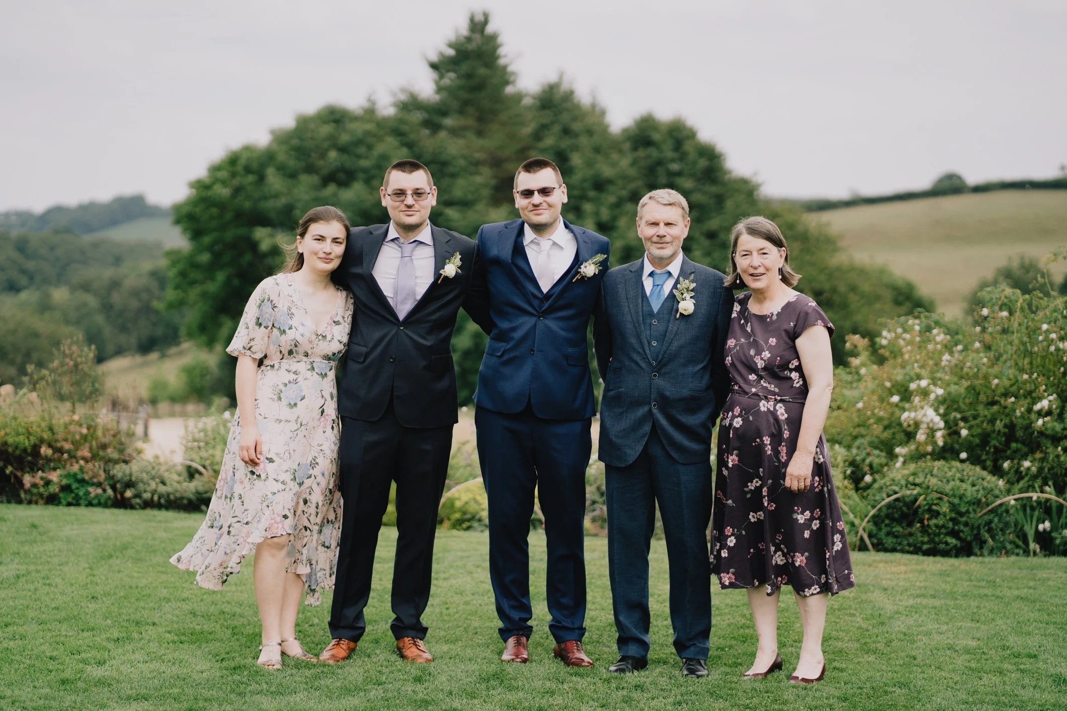 Family groups standing outdoors on a grassy field in front of trees and rolling hills, dressed in formal and semi-formal clothing, smiling, at a special occasion.