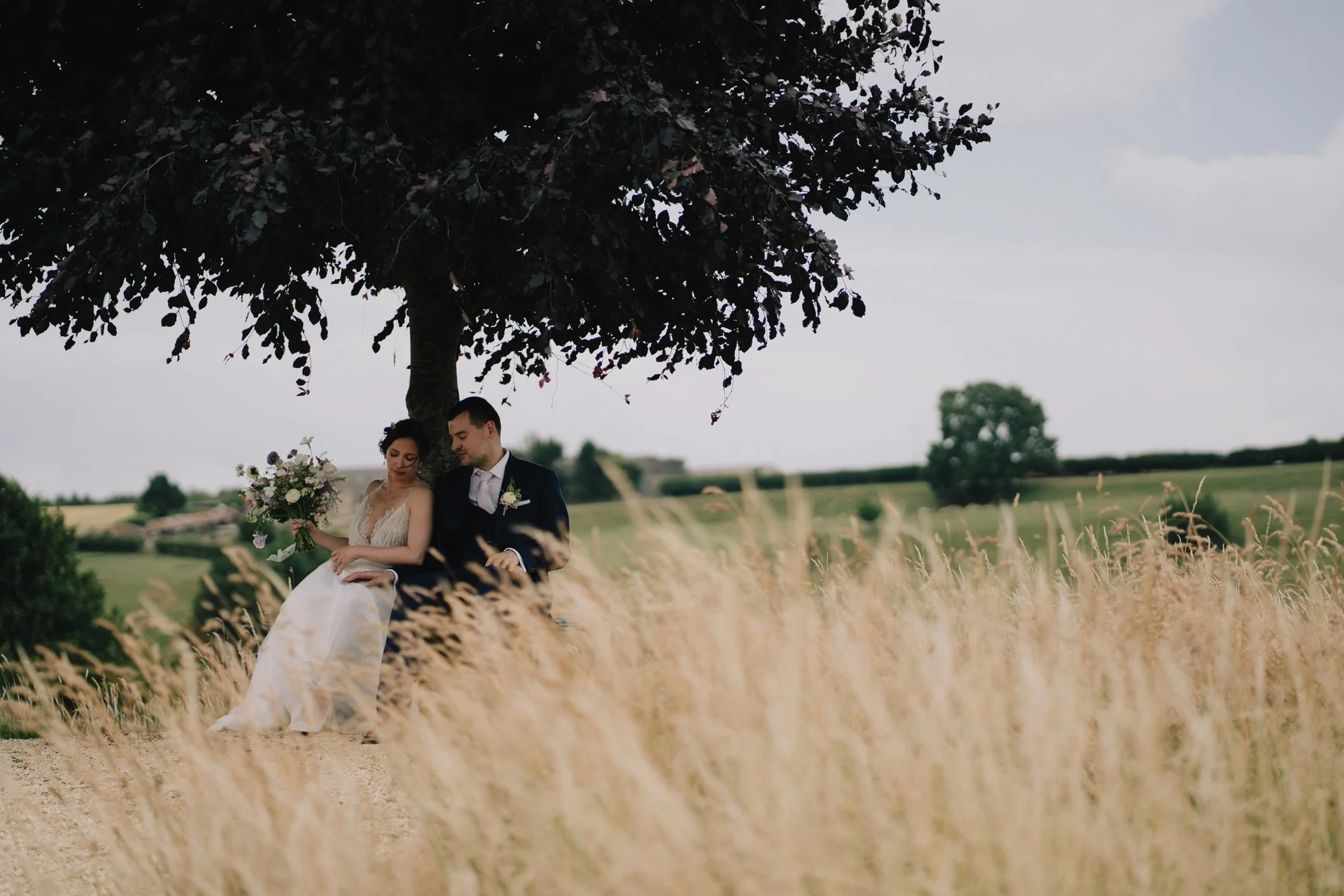 Bride and groom sitting together on a bench beside the hillside at Kingscote Barn