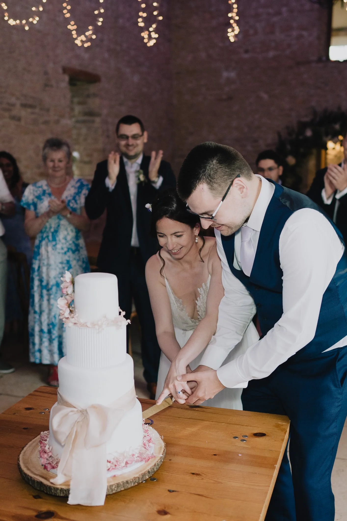 A bride and groom cutting a wedding cake together during their reception, with friends and family clapping in the background.