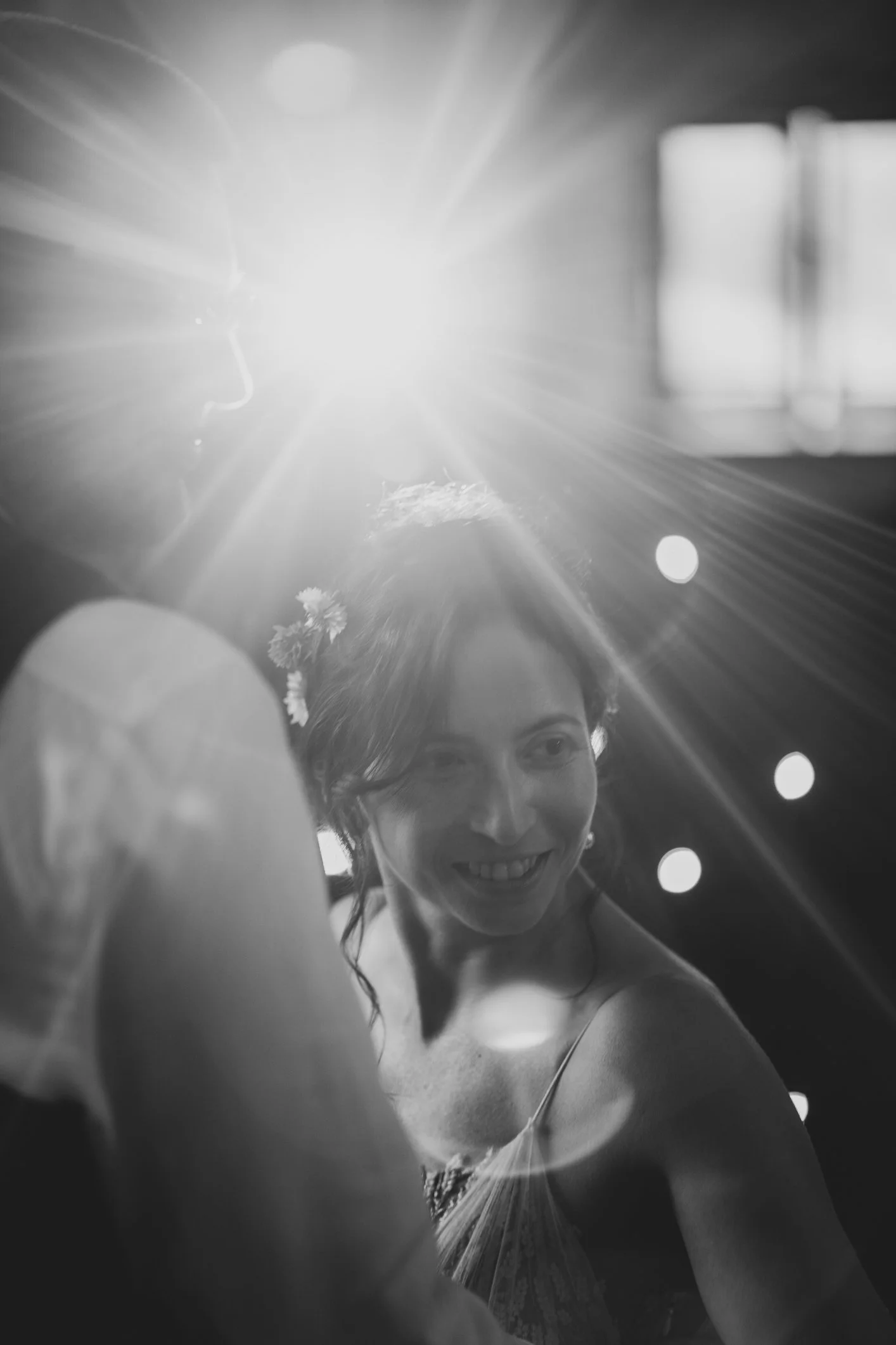 A woman smiling with flowers in her hair, illuminated by a bright light source behind her, creating a lens flare effect.