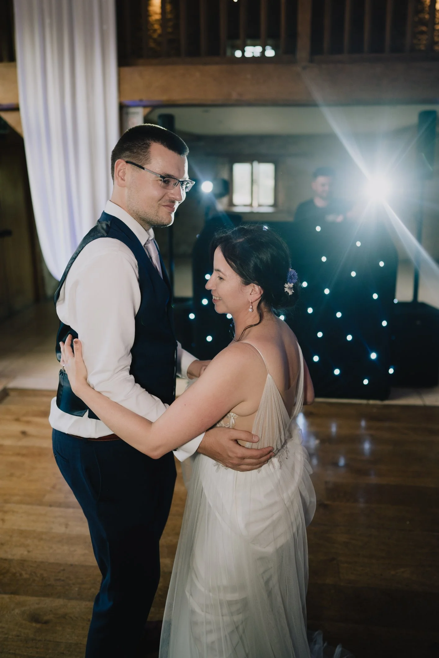 A couple dancing at a wedding reception, smiling and looking at each other, with a DJ setup in the background and natural light coming through a window.