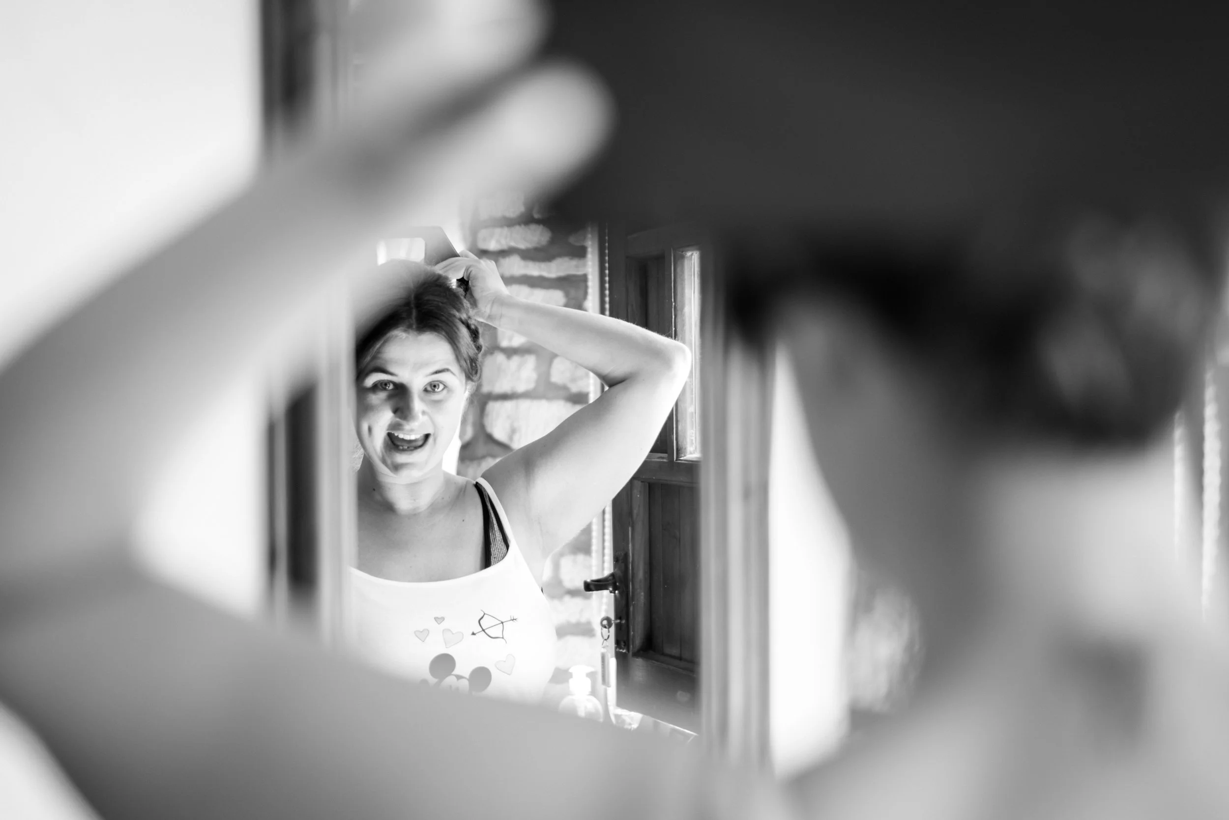 Black and white photo of a woman looking into a mirror, lifting her hair, with a playful or surprised expression, captured through a frame of a person or object in the foreground during prep at Great Tythe Barn.