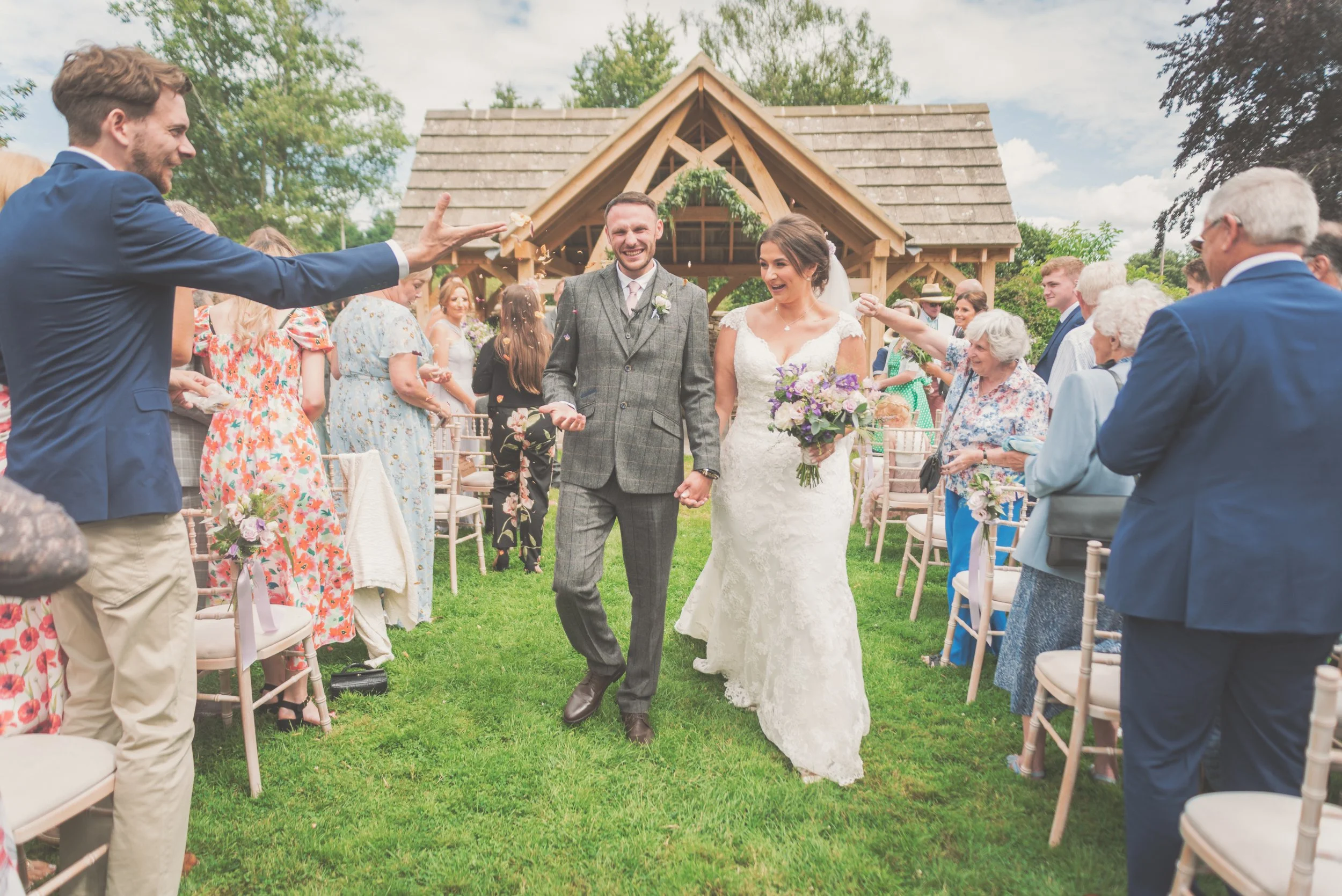 Bride and groom walking hand in hand through outdoor wedding aisle, smiling, surrounded by guests throwing confetti, with a wooden pavilion in the background taken by Great Tythe Barn wedding photographer.
