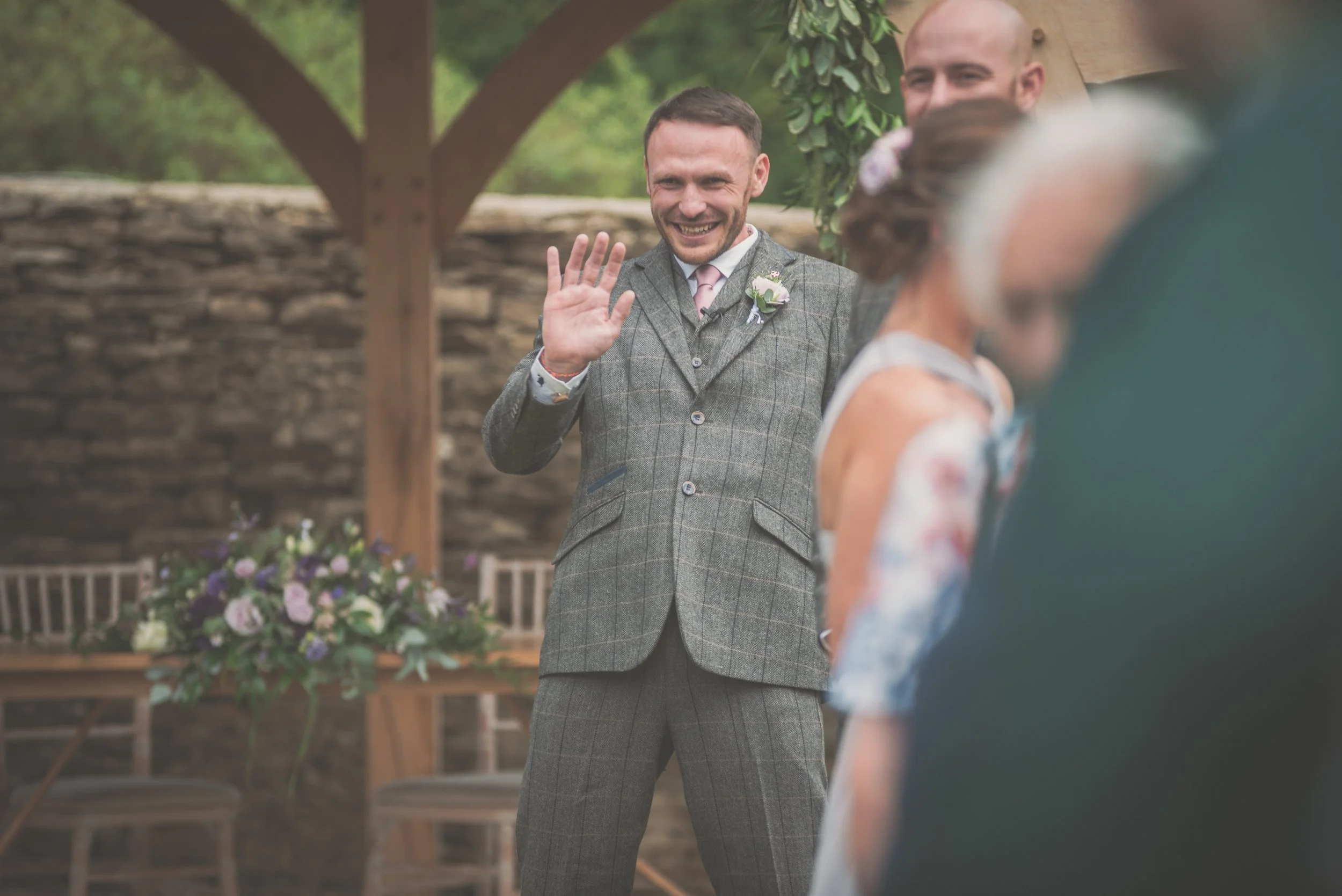 A groom in a gray plaid suit smiling and waving at an outdoor wedding ceremony. Behind him, a blurred bride, and another man are visible, with a wooden arch and flower arrangement in the background at Great Tythe Barn.