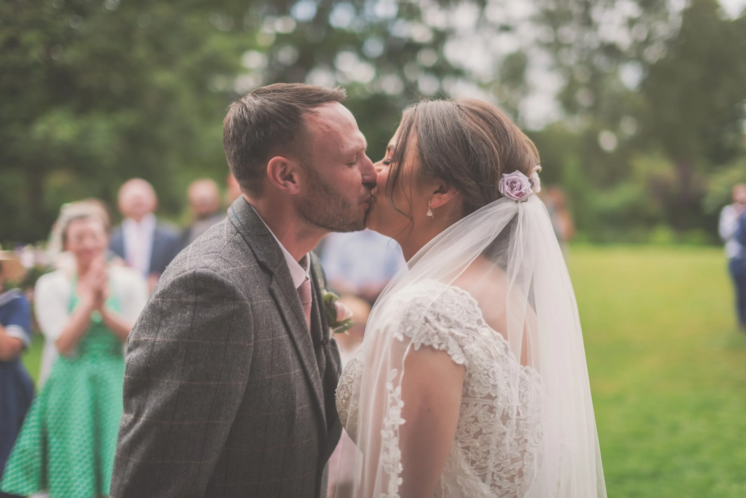 A bride and groom kiss during their outdoor wedding ceremony with guests in the background on a beautiful day taken by Great Tythe Barn Wedding Photographer