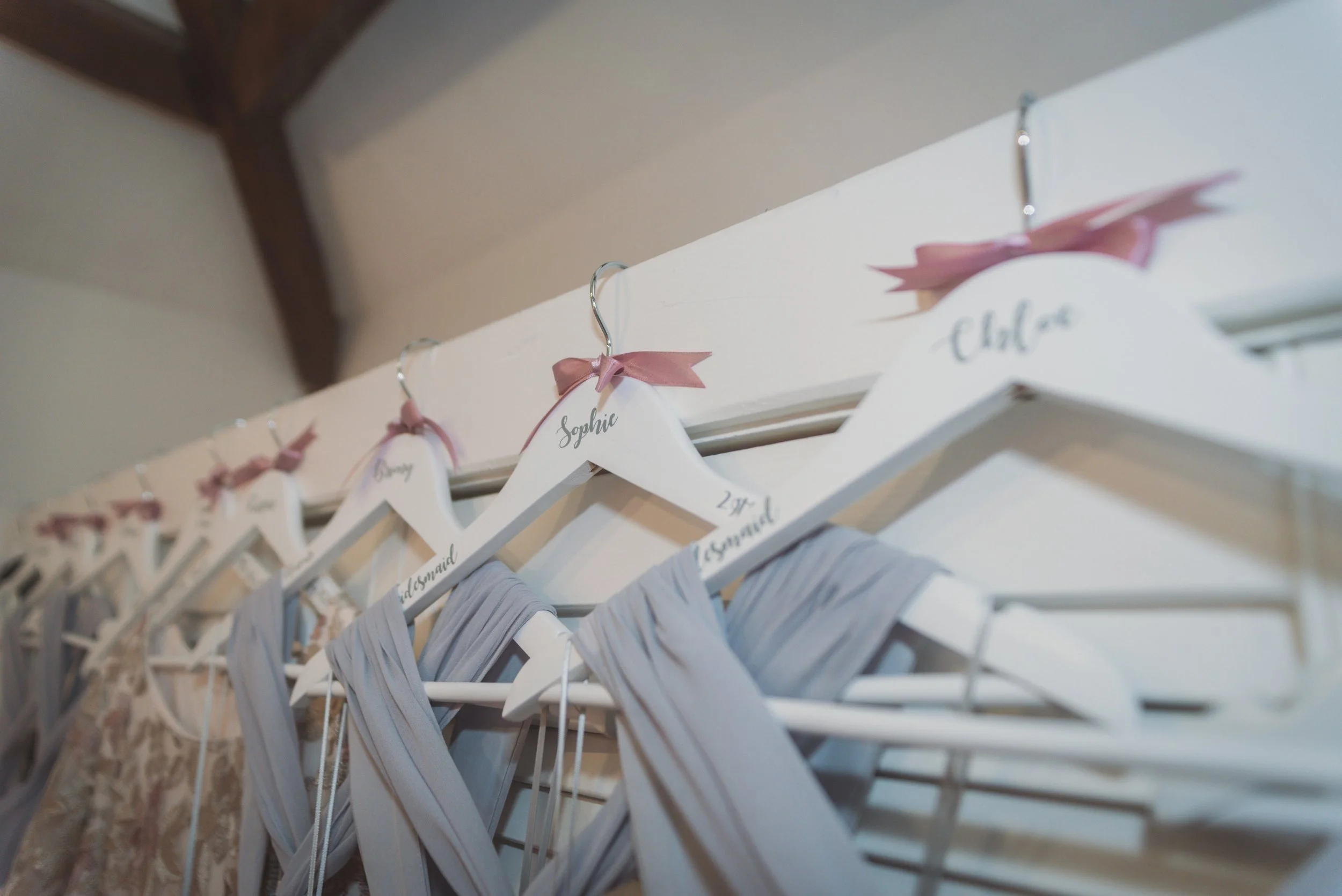 Hangers with lace and gray fabric clothing, each with a pink ribbon bow and a name tag, hanging on a white wooden rack, with one tag reading 'Sophie'.