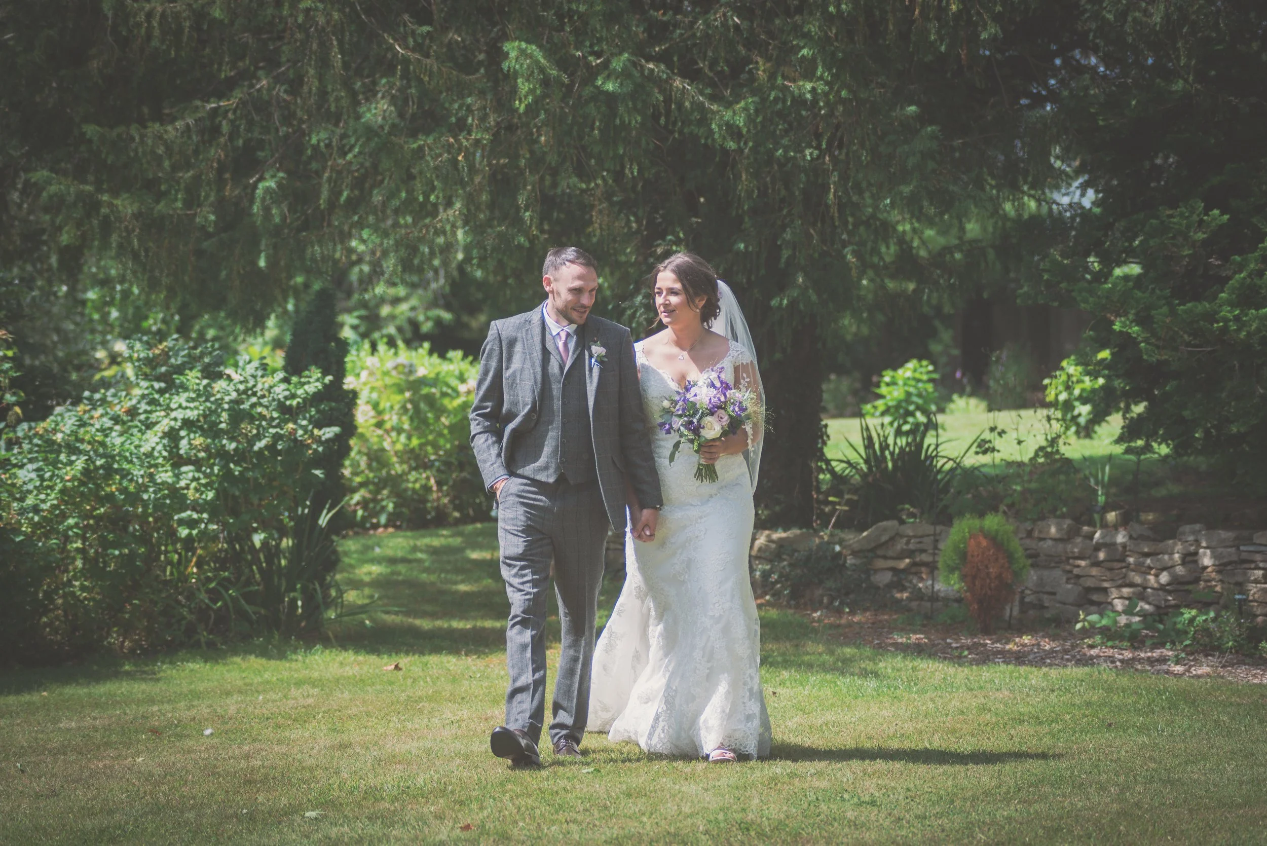 A newlywed couple walking outdoors in a garden, with the bride holding a bouquet of purple and white flowers, both smiling and dressed in wedding attire after their wedding at Great Tythe Barn.