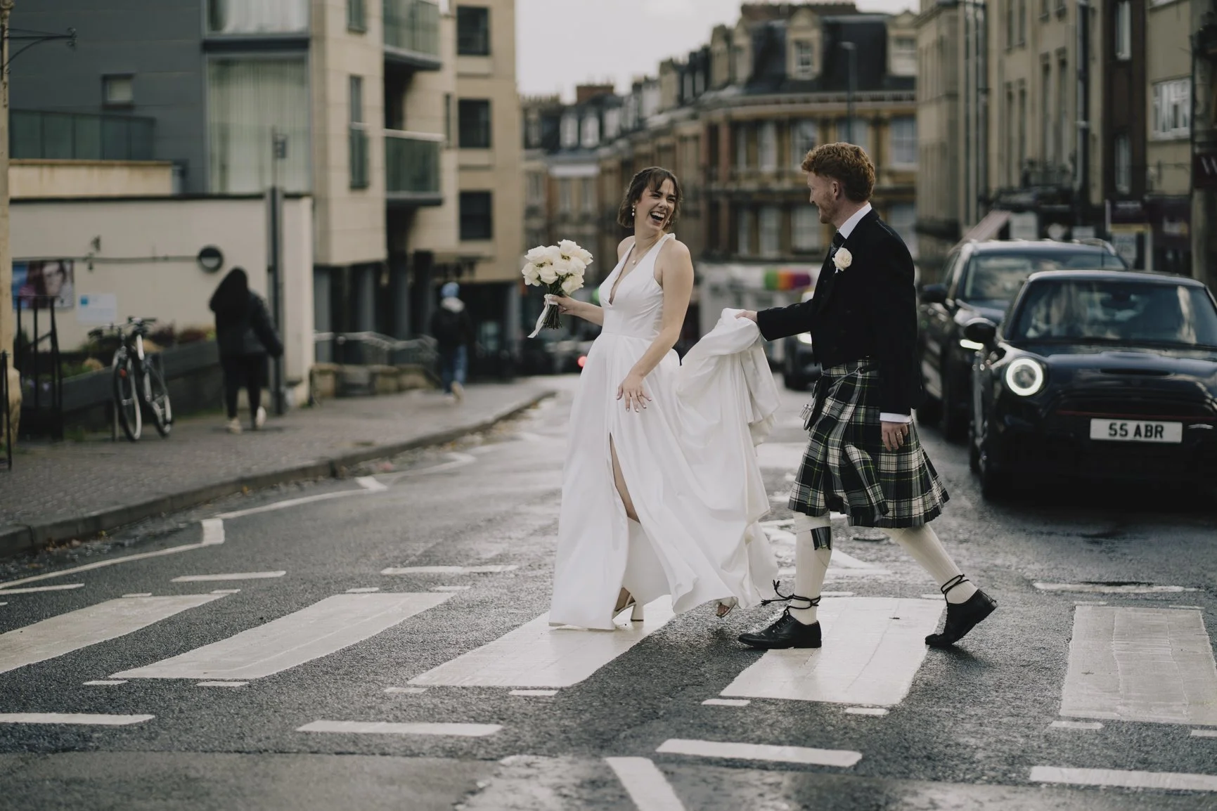 A newly married couple in wedding attire crossing a city street on a pedestrian crosswalk in Clifton, Bristol, sharing a joyful moment.