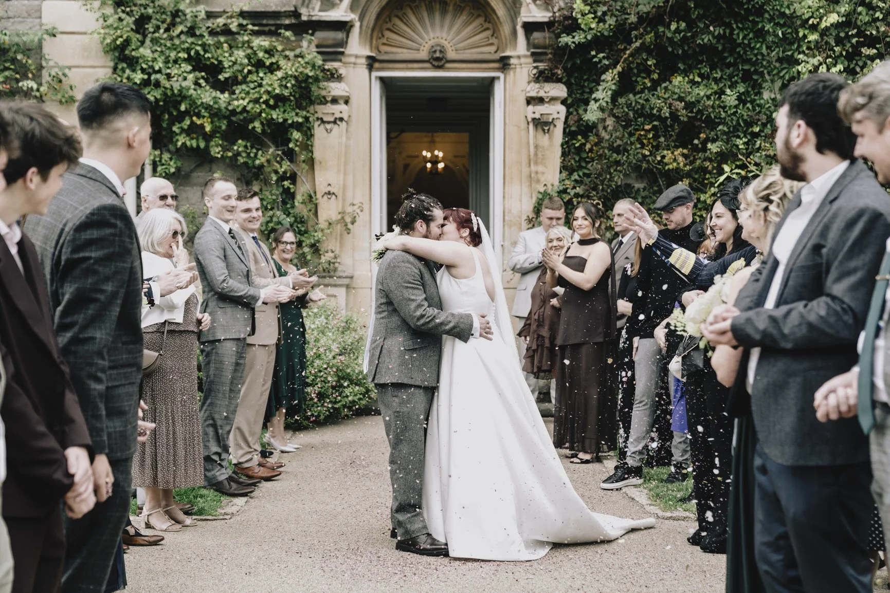 A newlywed couple shares a kiss at their wedding outside a historic building, surrounded by friends and family celebrating with confetti in Clevedon.