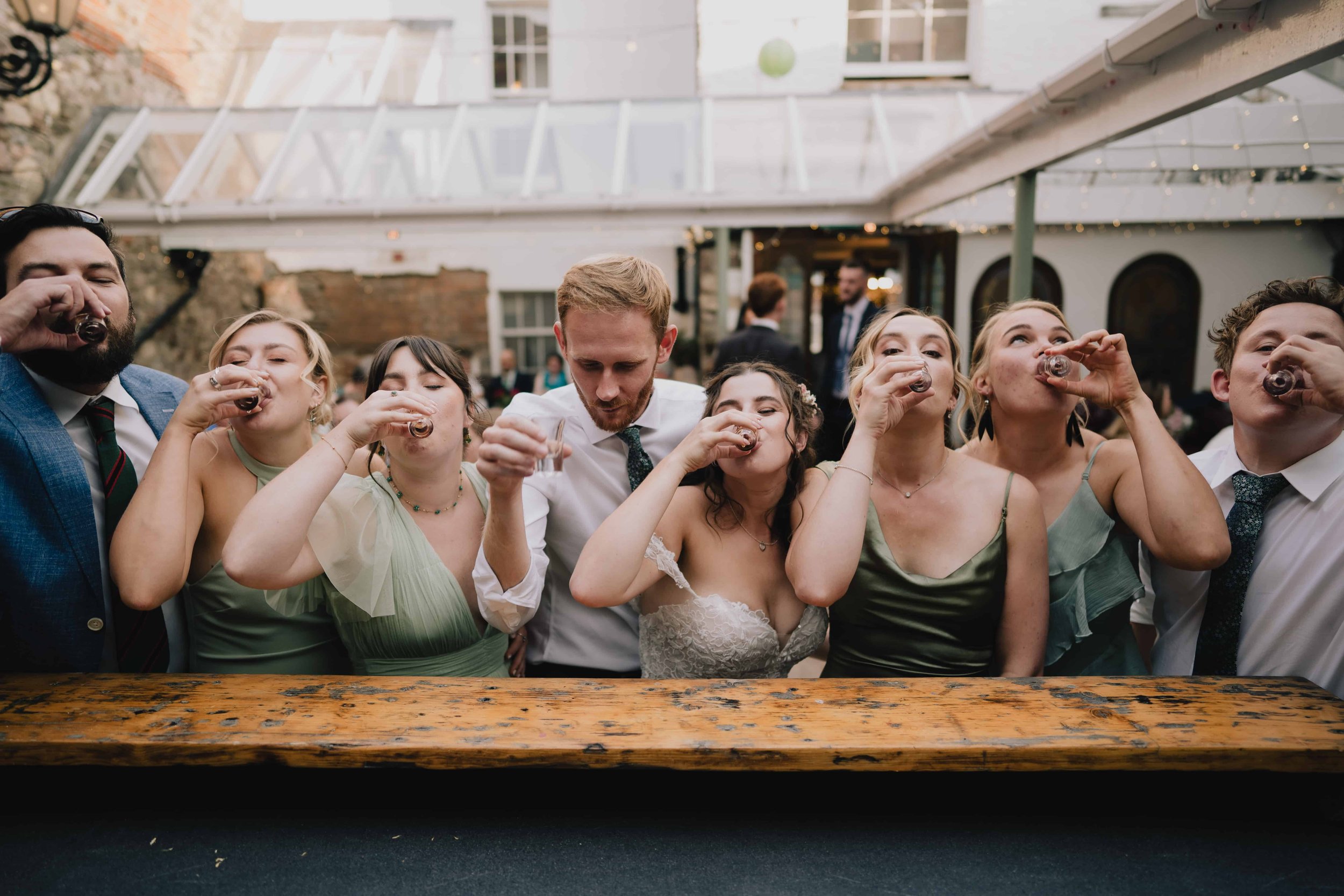 Group of people at a social event, toasting with shot glasses, outdoors under a glass roof.