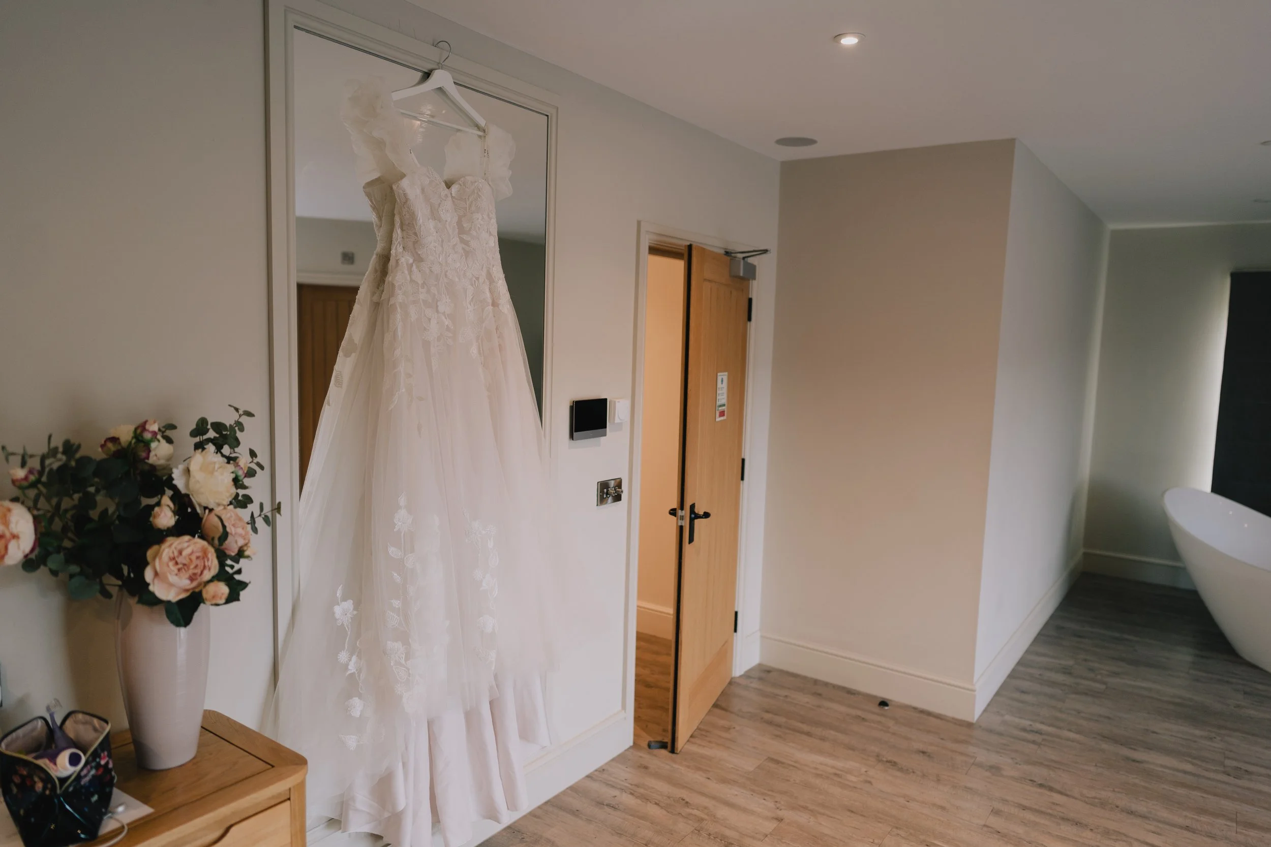 White wedding dress hanging on a hook in front of a mirror, next to a vase with pink and white flowers on a wooden table in a modern room.