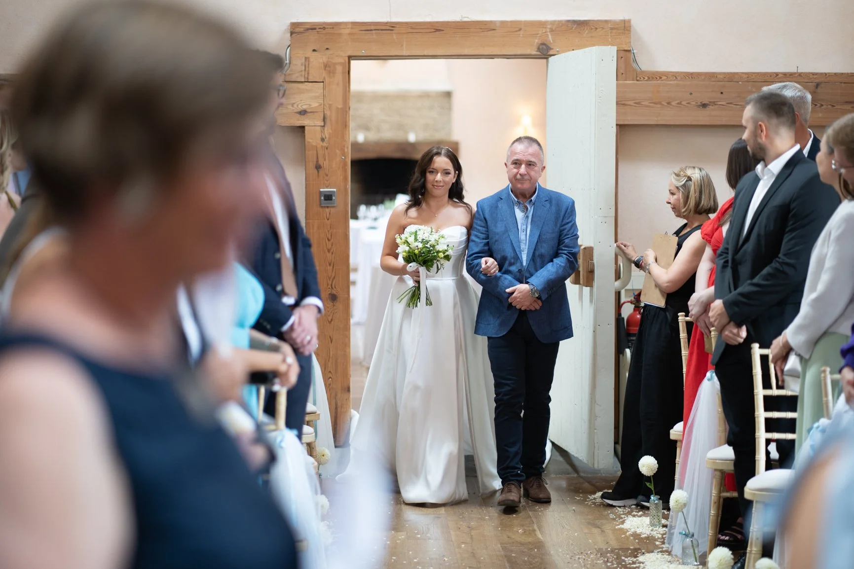 A bride walking down the aisle with her father at her wedding ceremony, surrounded by guests on either side.