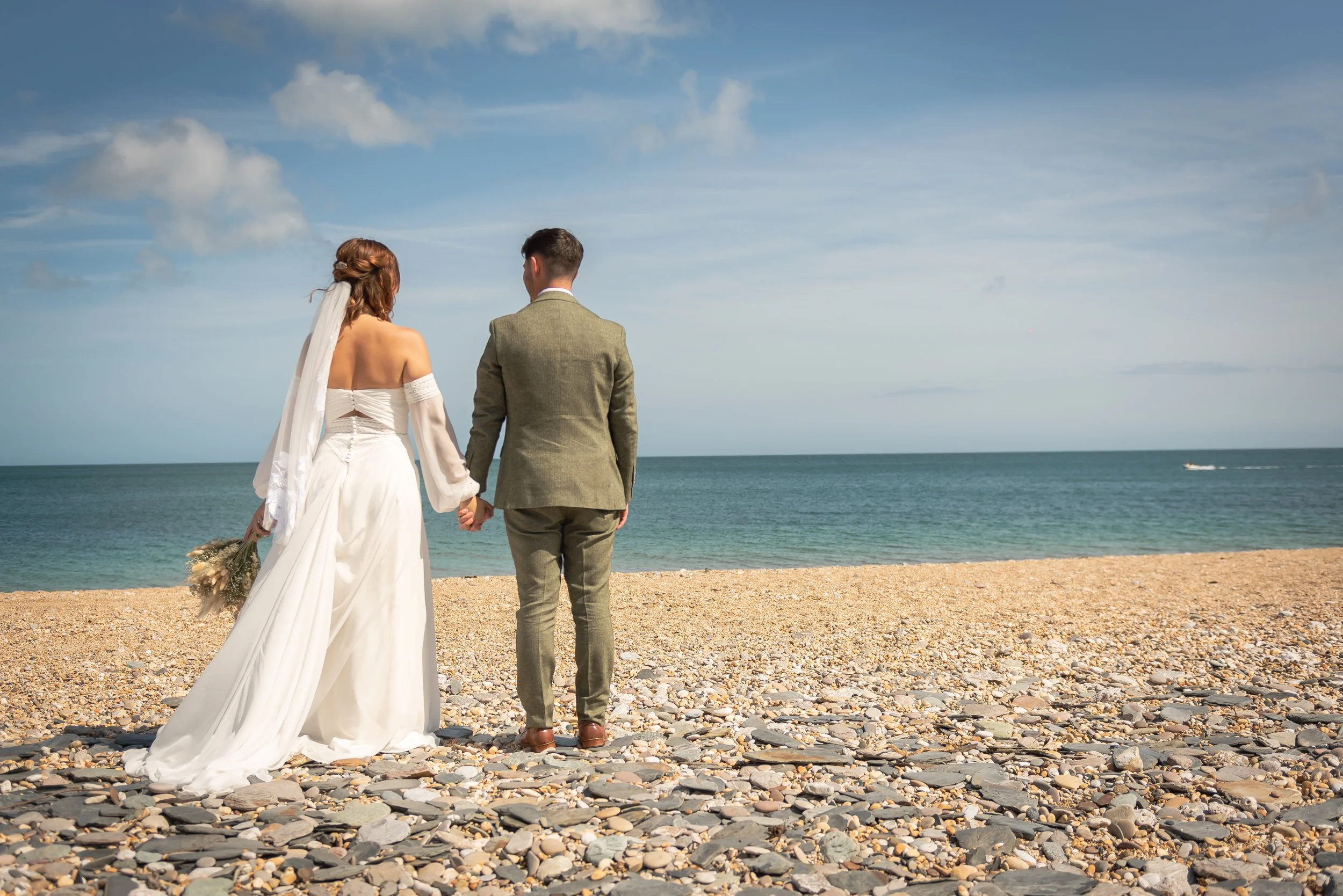 Bride and groom holding hands on a pebble beach, facing the ocean on a sunny day.