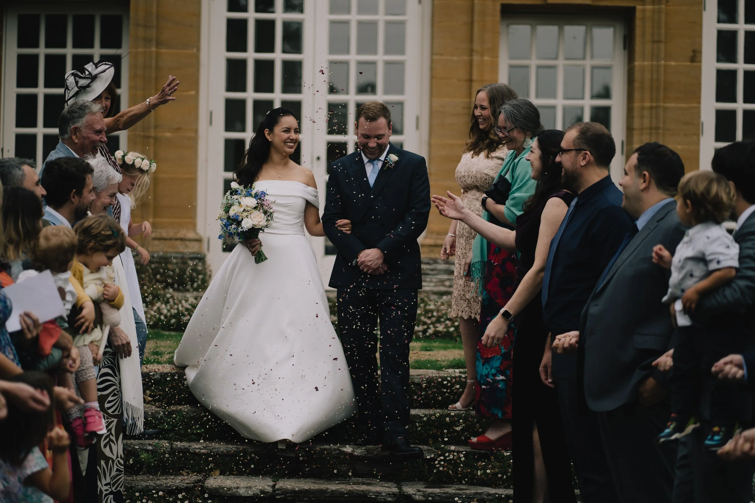 A bride and groom walking down the steps outside a building, surrounded by friends and family, celebrating their wedding with confetti and smiles.