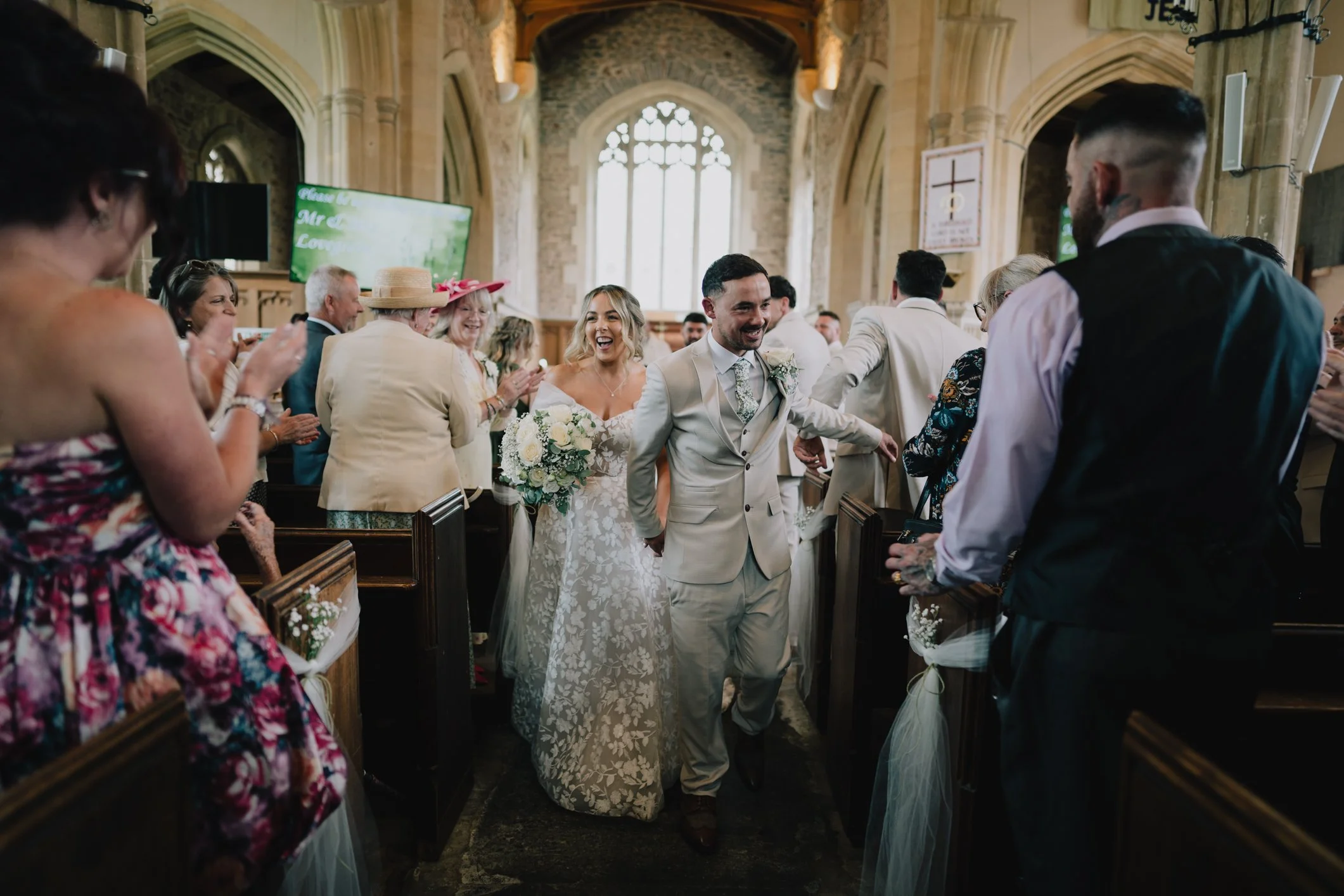 A bride and groom smiling as they walk down the aisle of a church after their wedding ceremony, surrounded by wedding guests clapping and smiling.