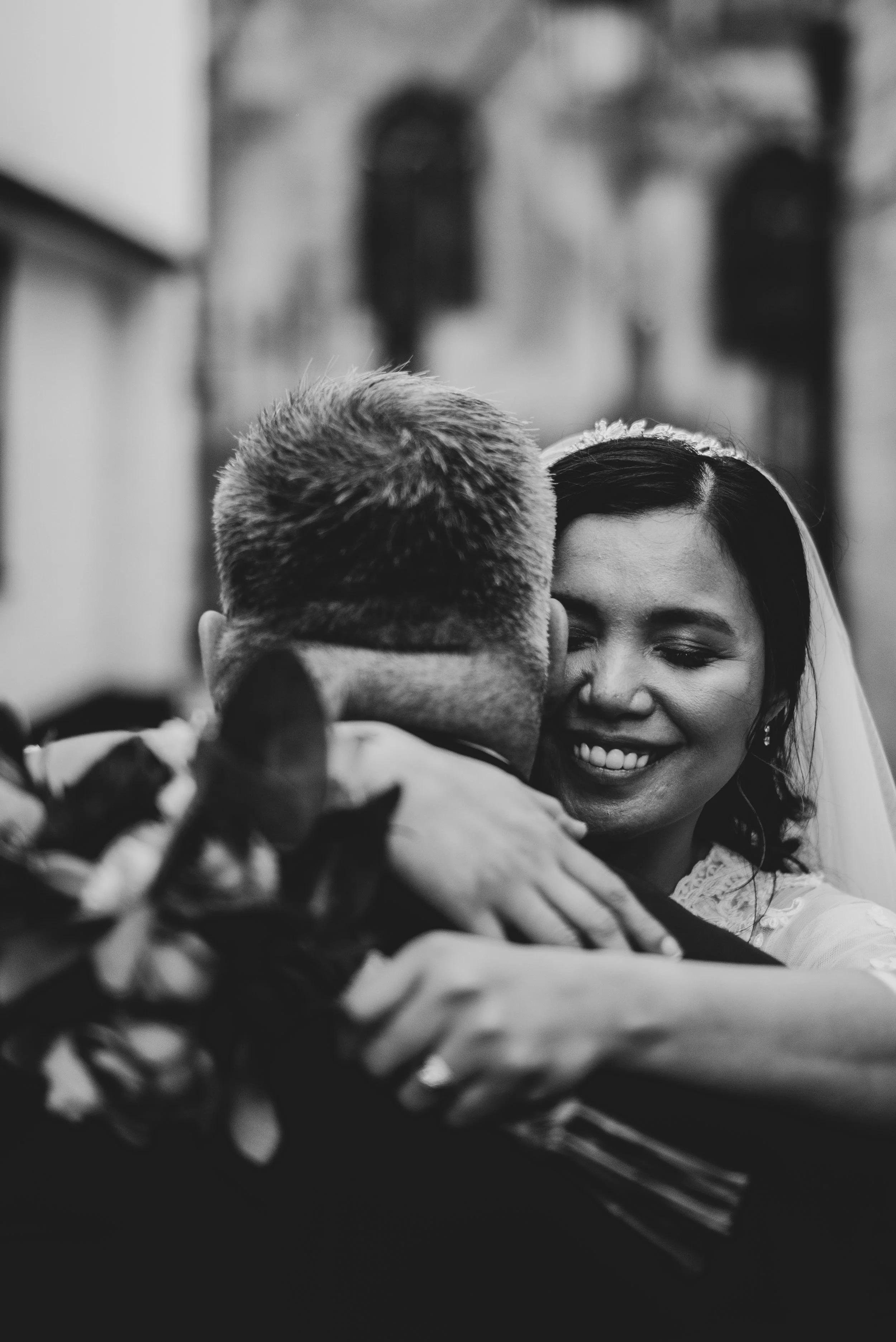 Black and white photo of a woman smiling and hugging a man, seen from behind, with flowers in the foreground.