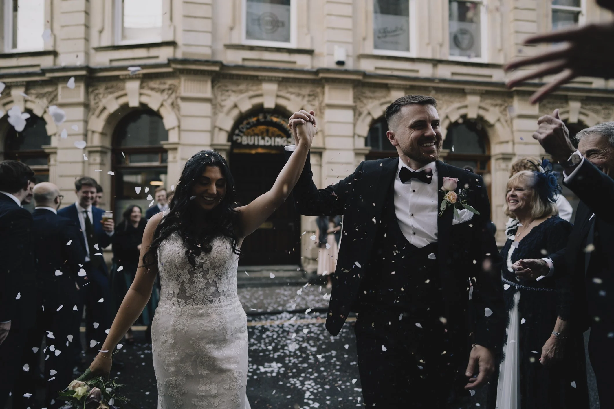 A newly married couple holding hands and smiling as they walk through confetti with wedding guests around them in front of a building in Bristol.