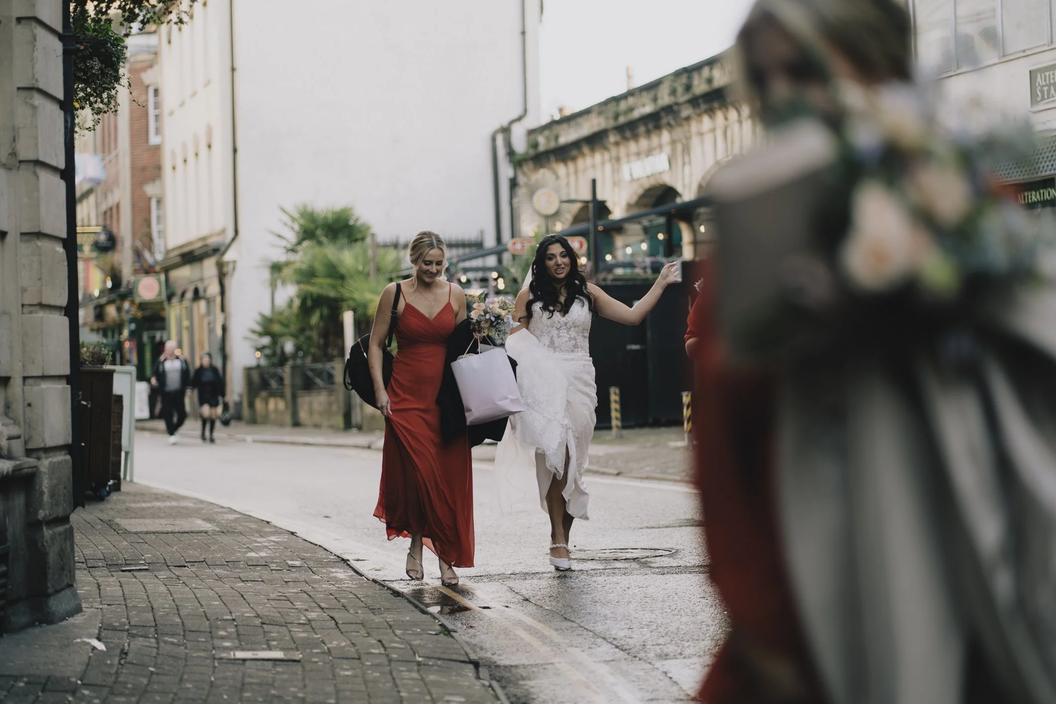 Two women, one in a red dress and the other in a white wedding dress, walking on a city sidewalk holding flowers and a gift bag, with a blurred car in the foreground and a city street background in Bristol